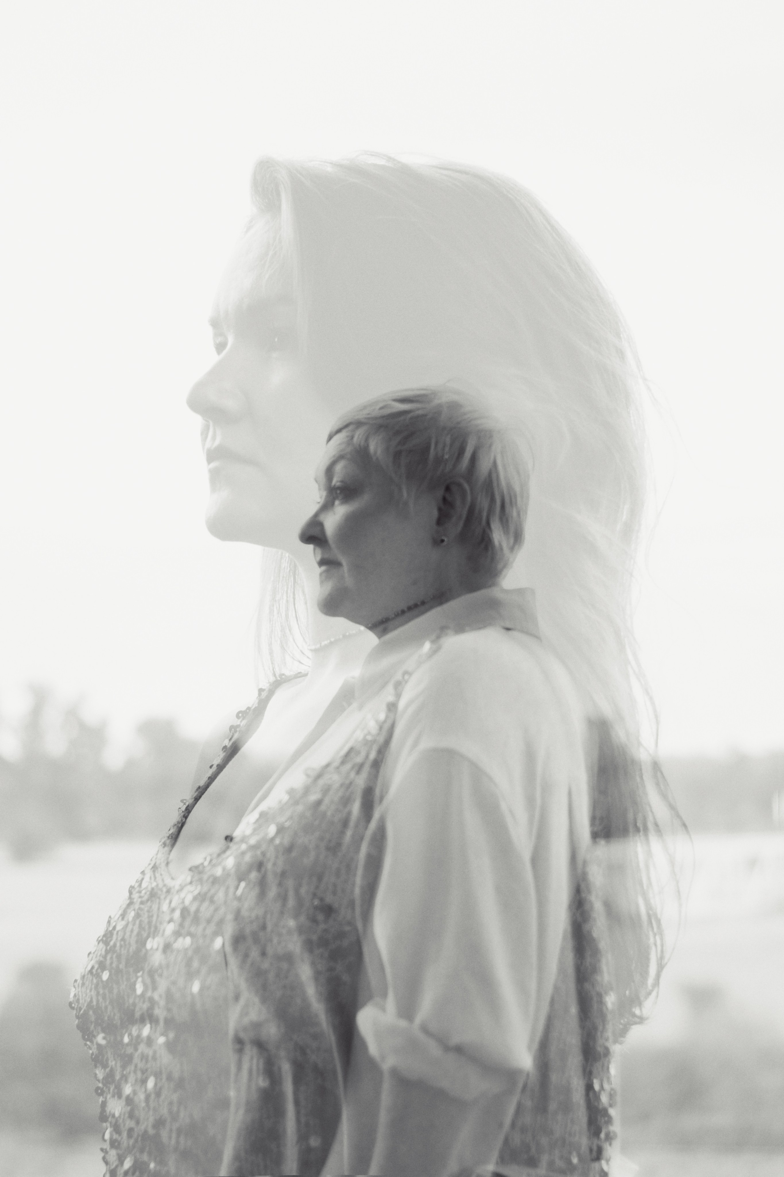 На фотографии изображены две женщины, стоящие спиной к камере. Одна из них взрослая, а другая молодая. Обе они выглядят элегантно и торжественно. Взрослая женщина одета в длинное платье с рукавами-фонариками и глубоким декольте, украшенное кружевной отделкой. Молодая женщина также в длинном платье с прозрачными рукавами и украшениями, напоминающими кристаллы. Волосы обеих женщин аккуратно уложены. На заднем плане видны водоем и деревья, что указывает на то, что они находятся на природе.