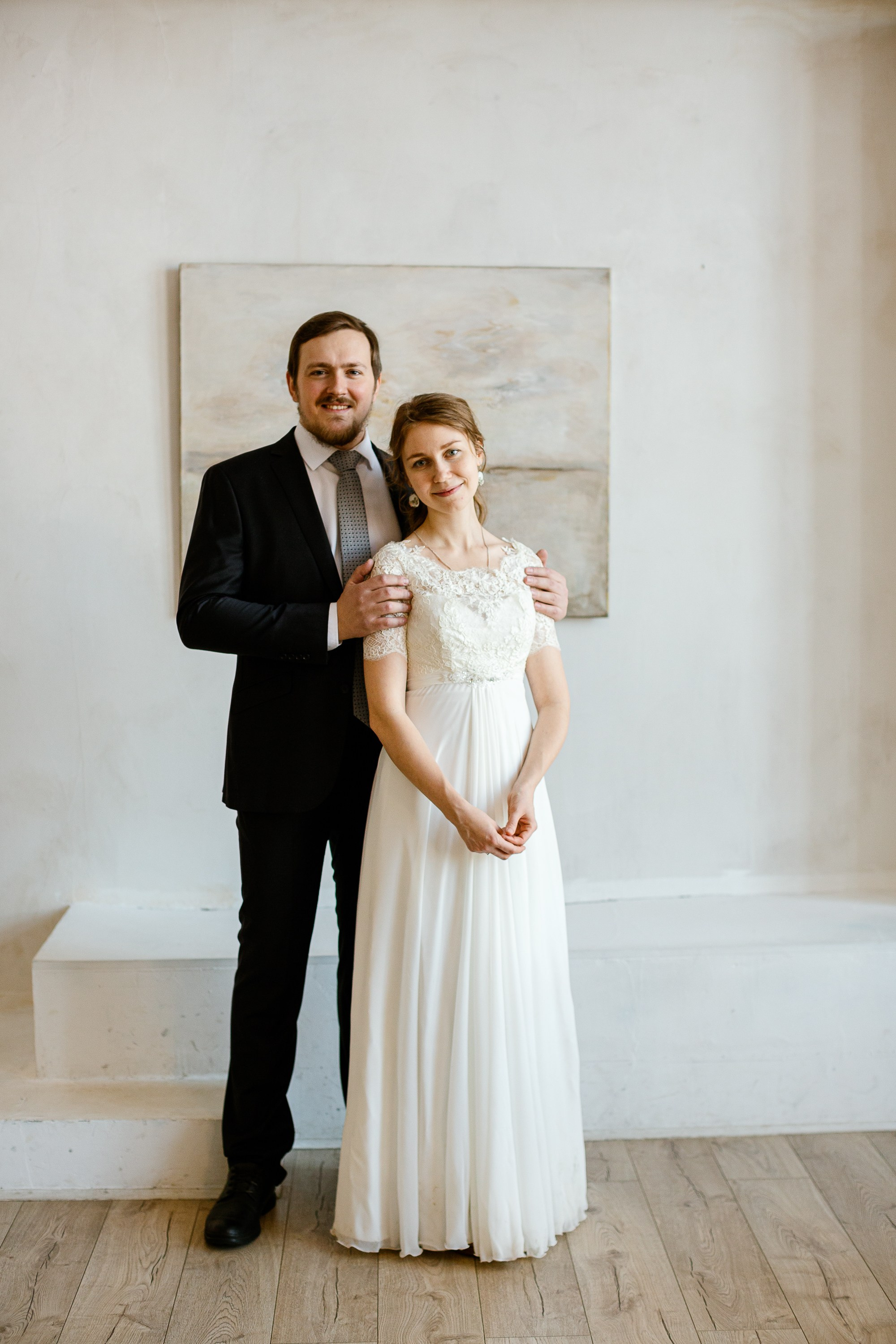 Жених и невеста в праздничной фотостудии. Жених в костюме, невеста в красивом белом платье. На фоне картина.