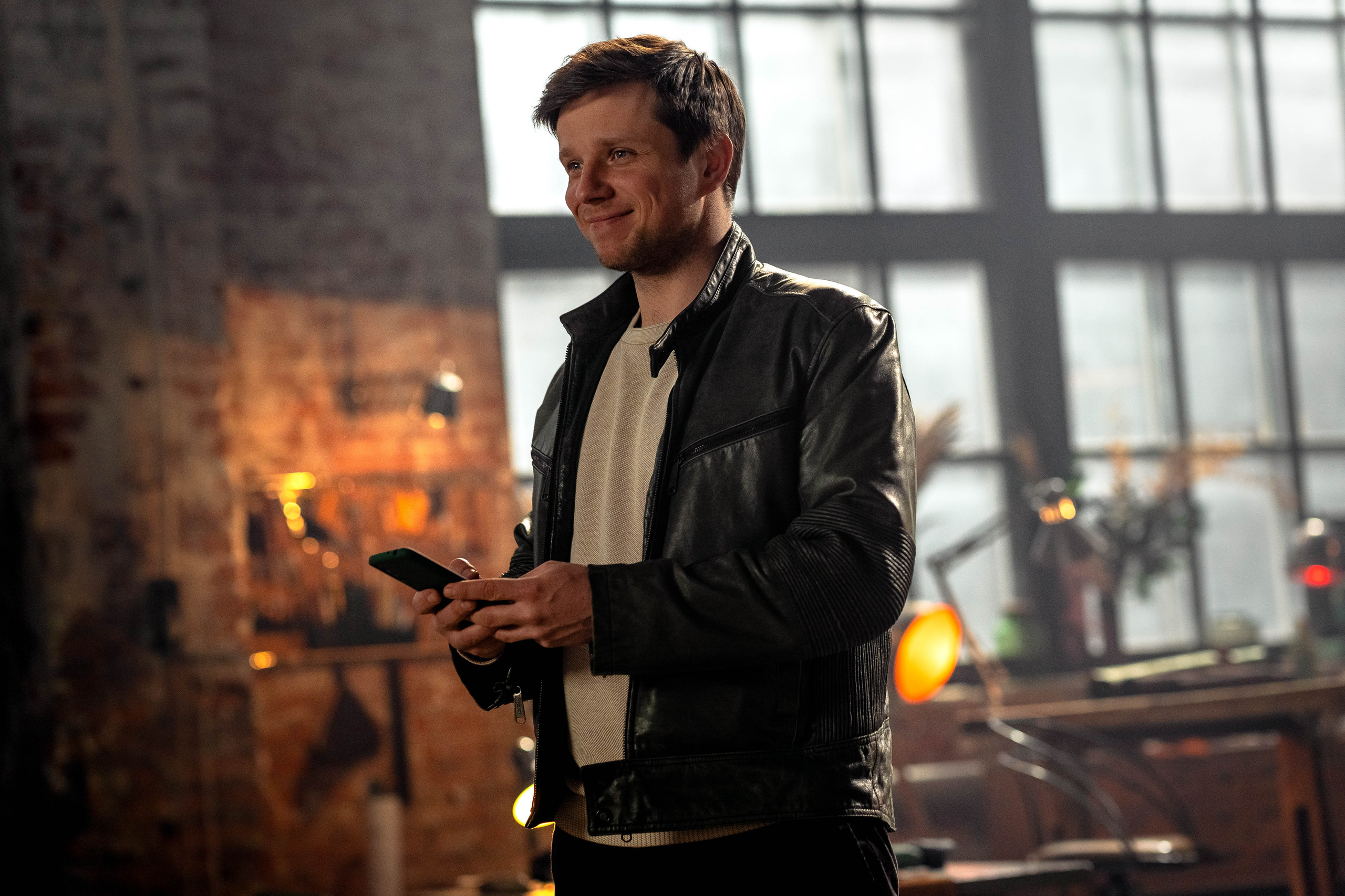 Молодой мужчина брюнет в зеленой куртке стоит с телефоном в руках в лофт-помещении