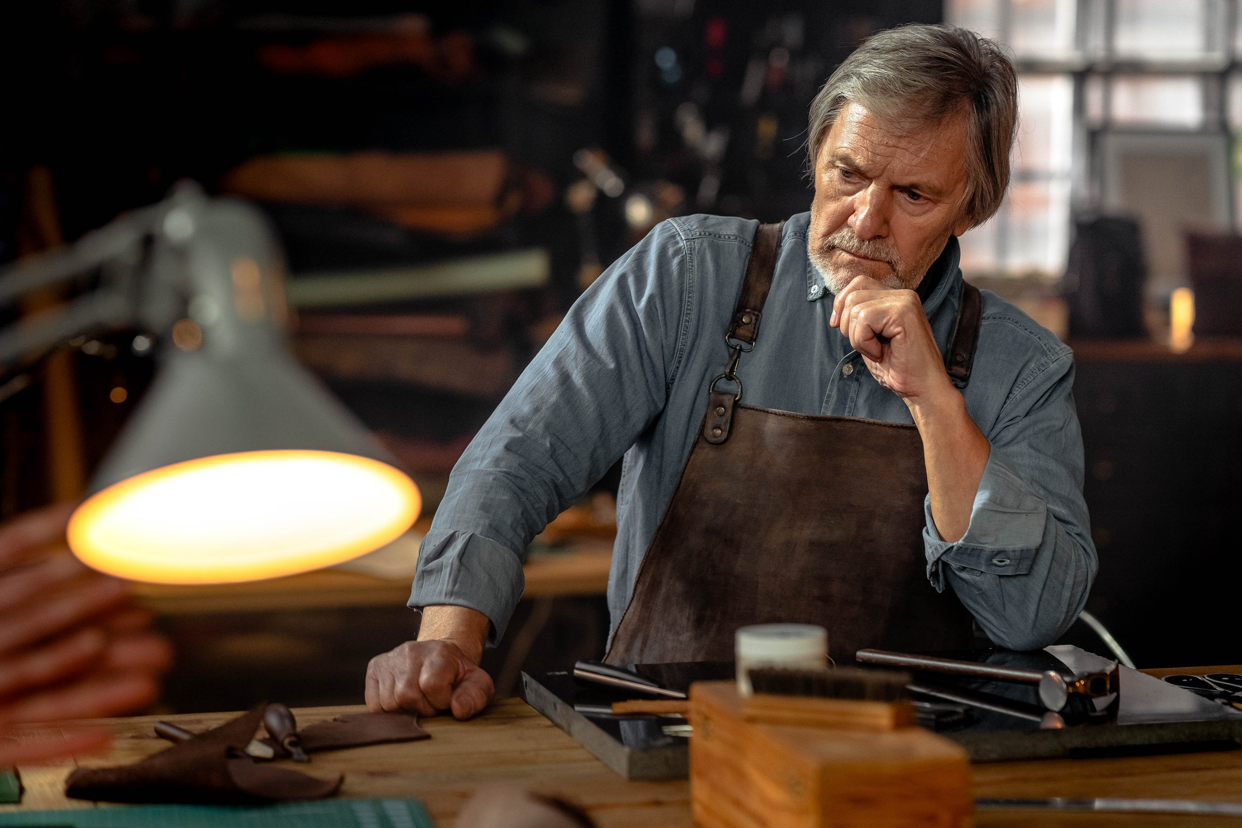 Пожилой седой мужчина в кожаном фартуке сидя за столом, задумчиво смотрит в сторону, подперев рукой подбородок