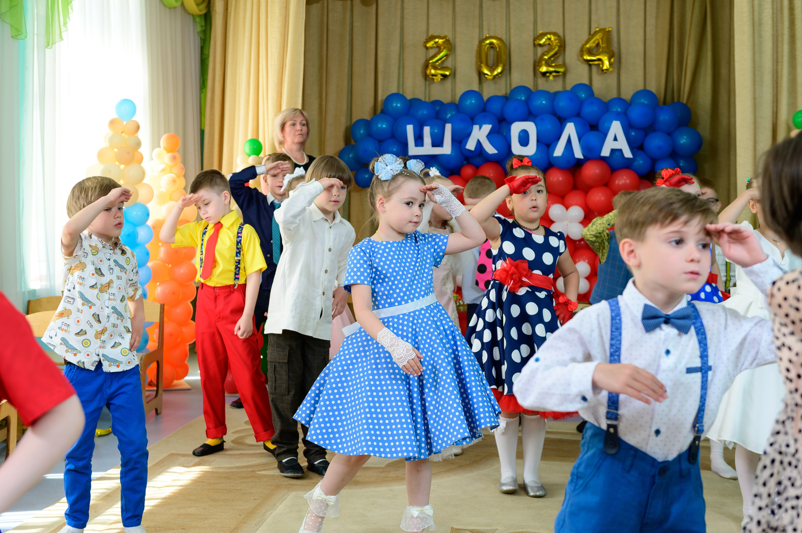 Дети танцуют на утреннике в детском саду. Девочка в голубом платье в центре зала.