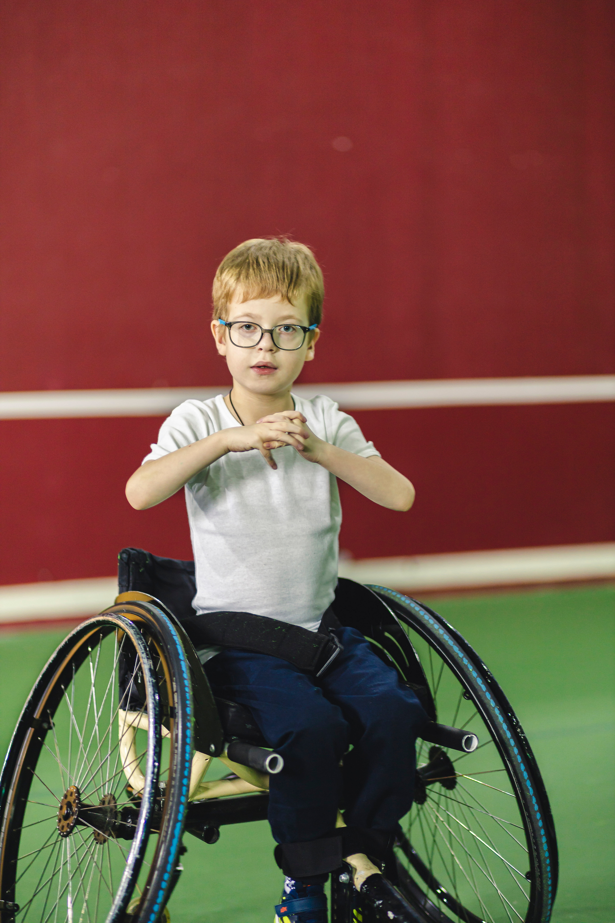 мальчик на коляске в очках и белой футболке на фоне красной стены и зеленого пола 