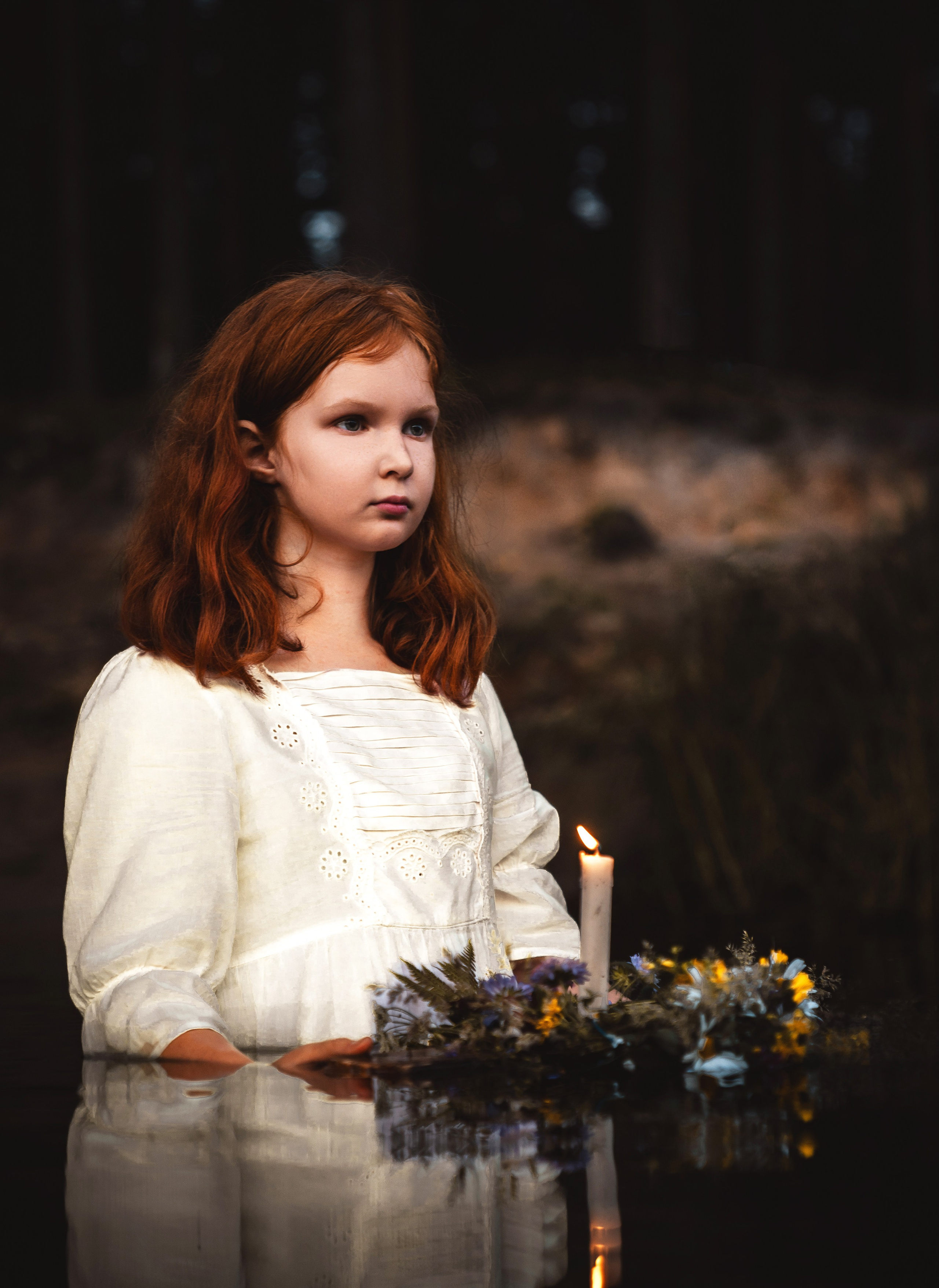 Рыжая девочка по щиколотку в лесной воде держит горящую свечу и венок из полевых цветов.