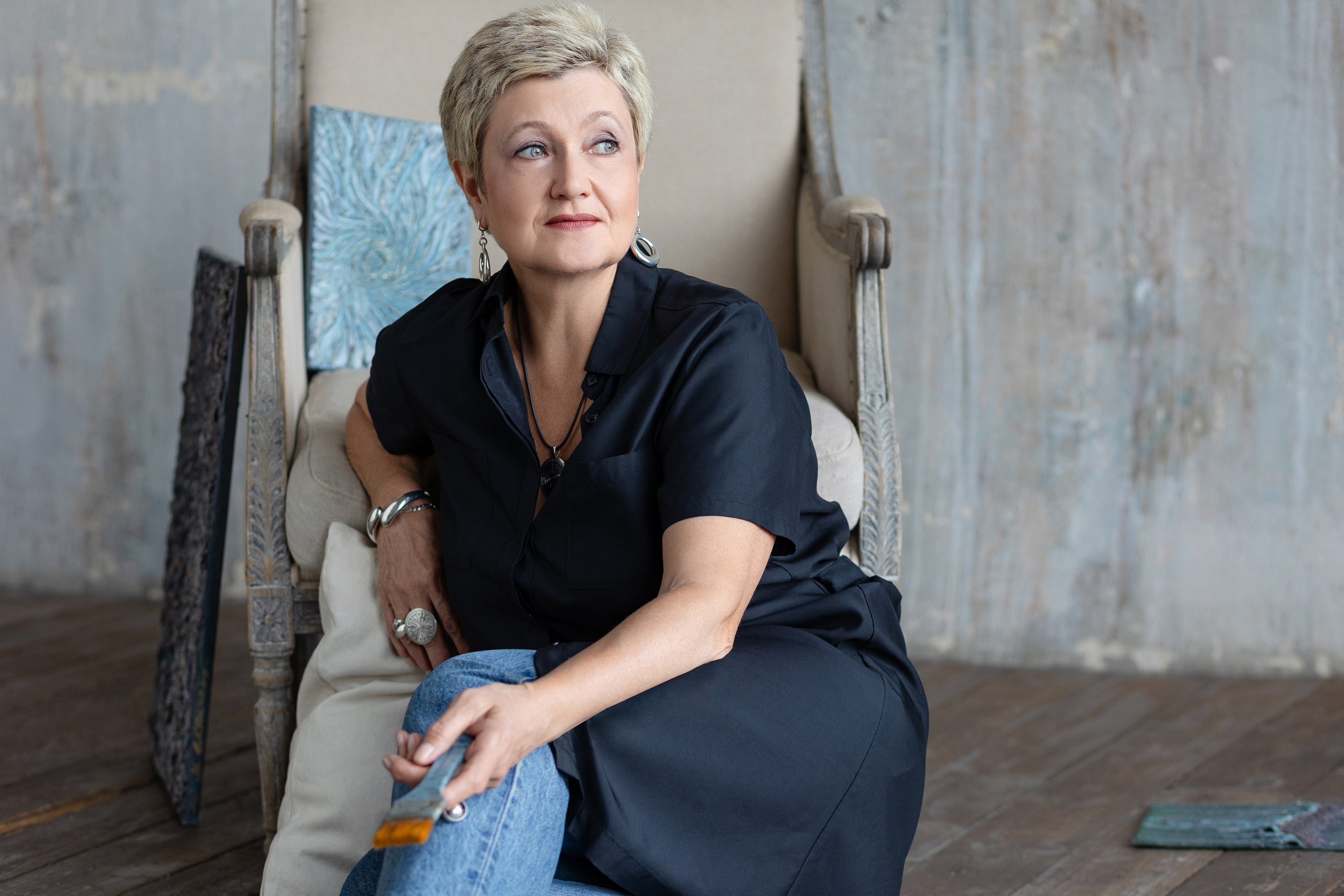 Женщина сидит возле кресла на полу, под рукой у нее подушки.