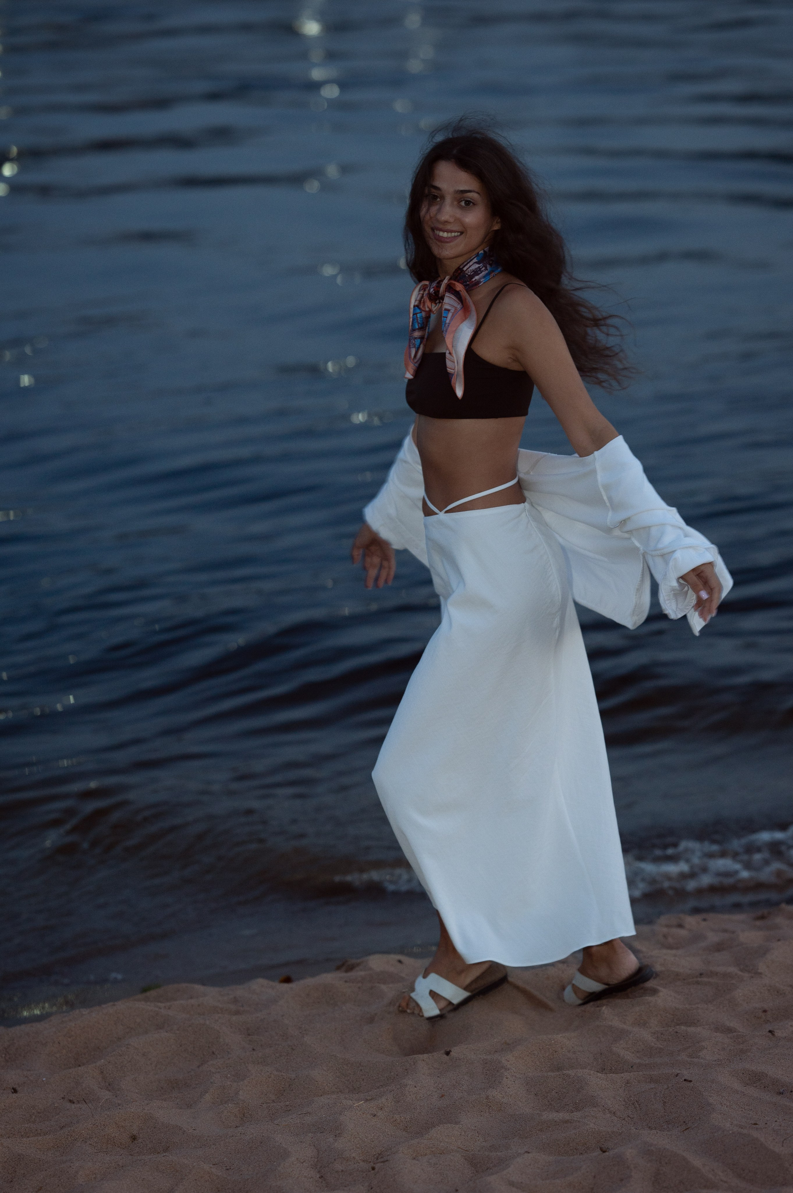 Красивая длинноволосая брюнетка со смуглой кожей в белой одежде идет по берегу реки, на шее у нее яркий платок