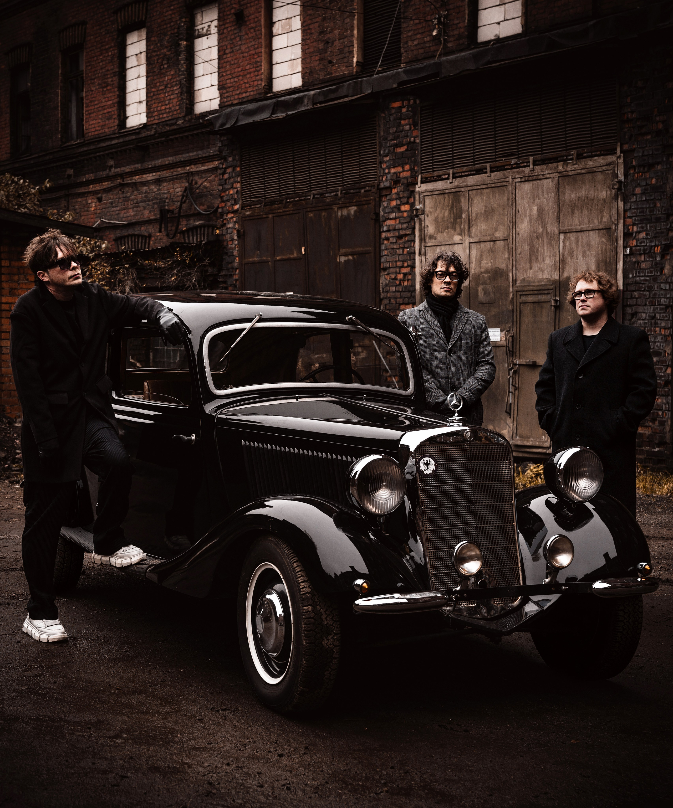 Радио Сатурн. Музыканты альтернативной группы в длинных чёрных пальто, очках и кедах позируют у винтажного чёрного автомобиля   Mercedes 30-х годов на фоне заброшенного кирпичного завода.