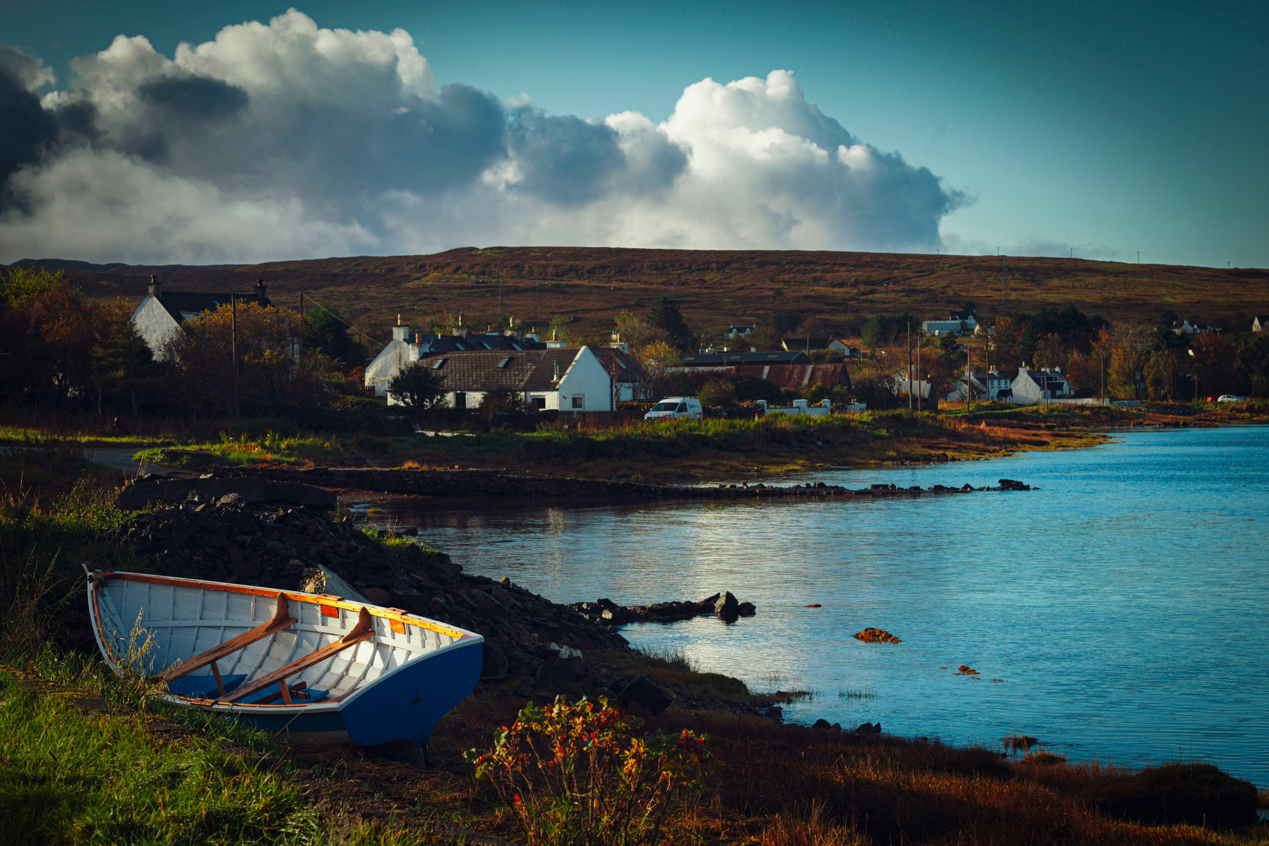 Белая деревянная лодка, стоящая на зеленом берегу у залива, с деревней белых домов и холмами на заднем плане под драматичным облачным небом.