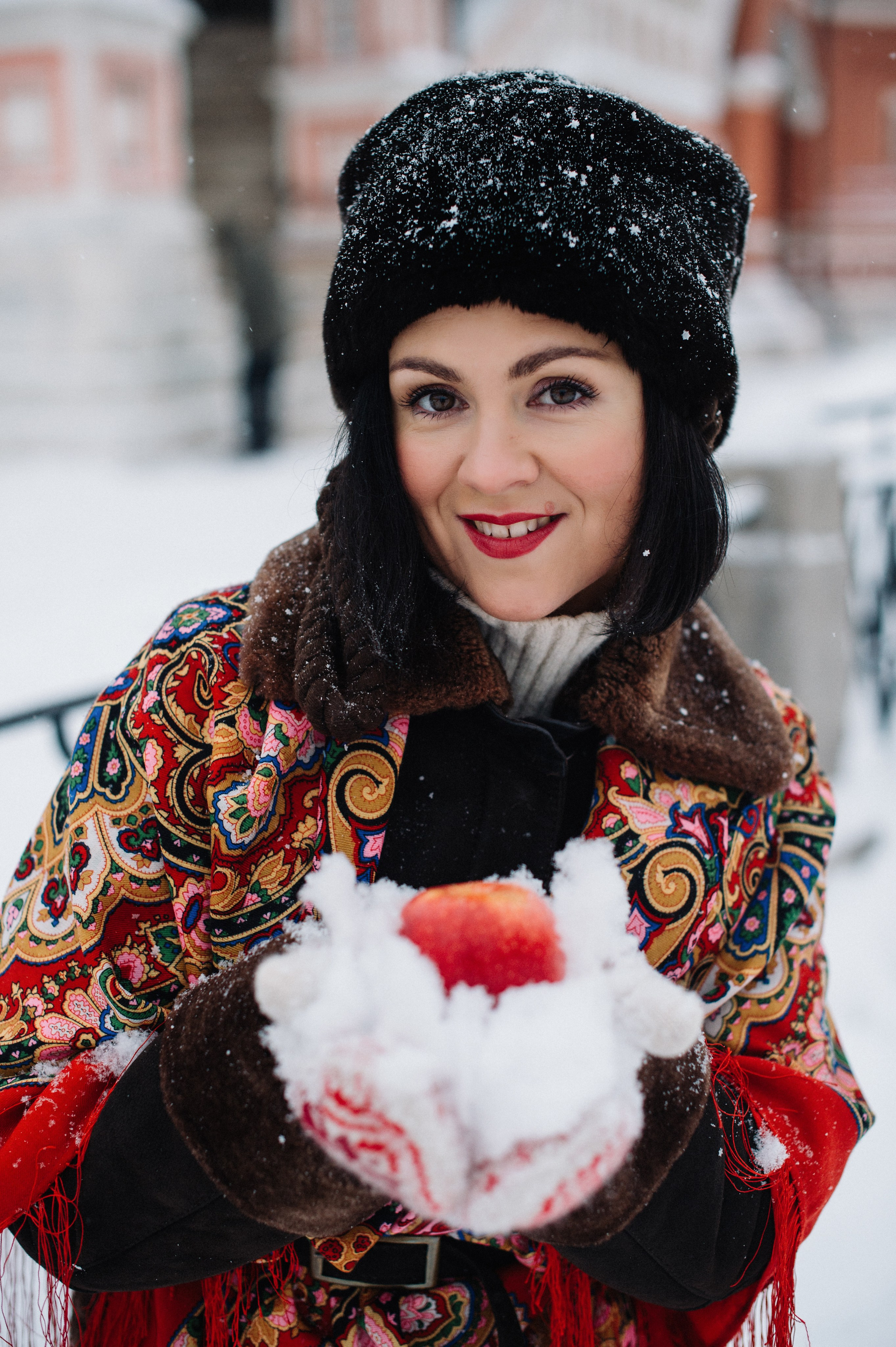 Девушка в красивом наряде в русском стиле с красным платком на плечах на Красной площади зимой. Она держит в руках в заснеженных варежках красные яблоки