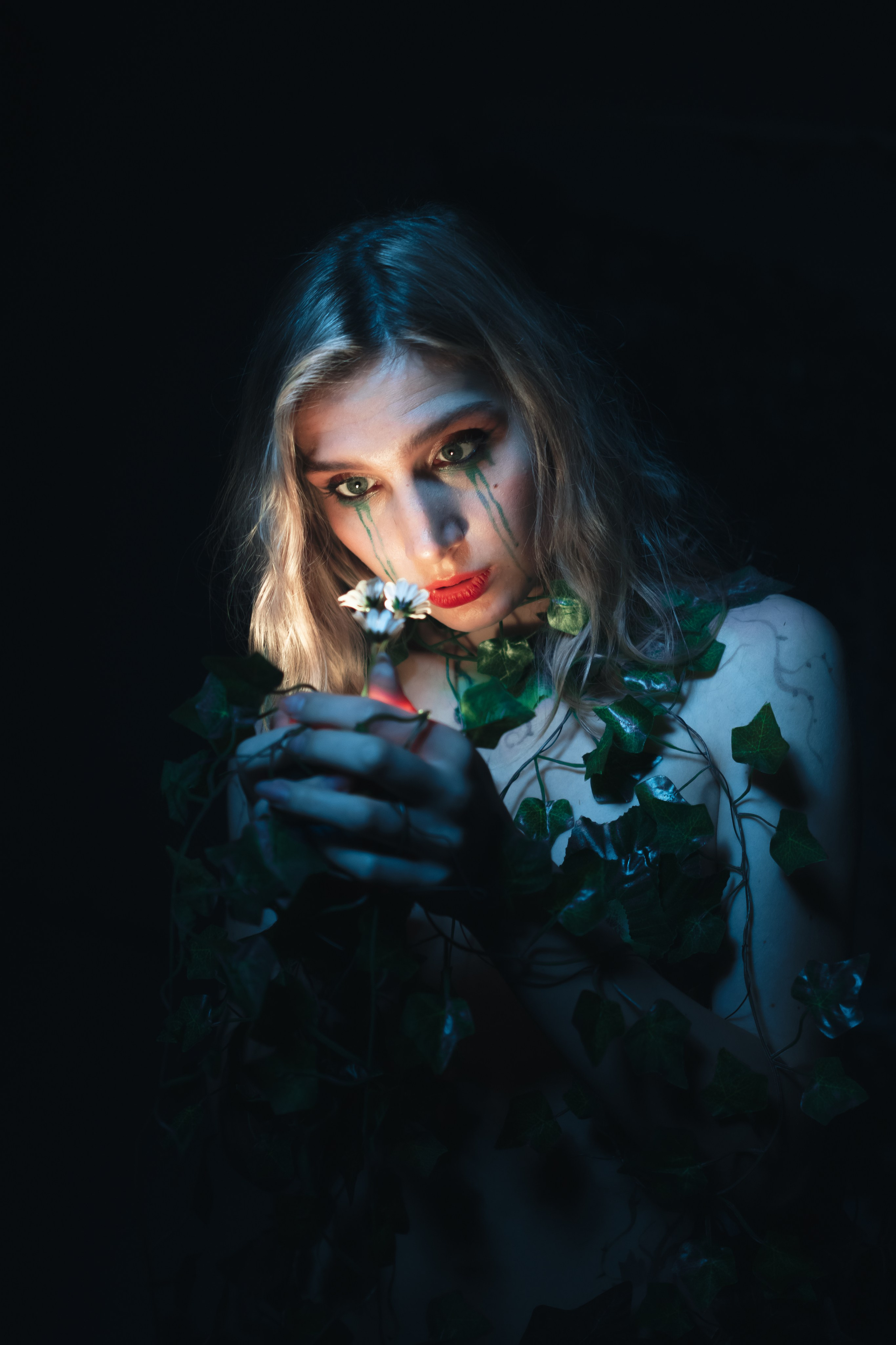 Художественная фотосессия в фотостудии во Владивостоке, девушка опутанная плющом с красными губами в синем свете смотрит на маленький цветочек