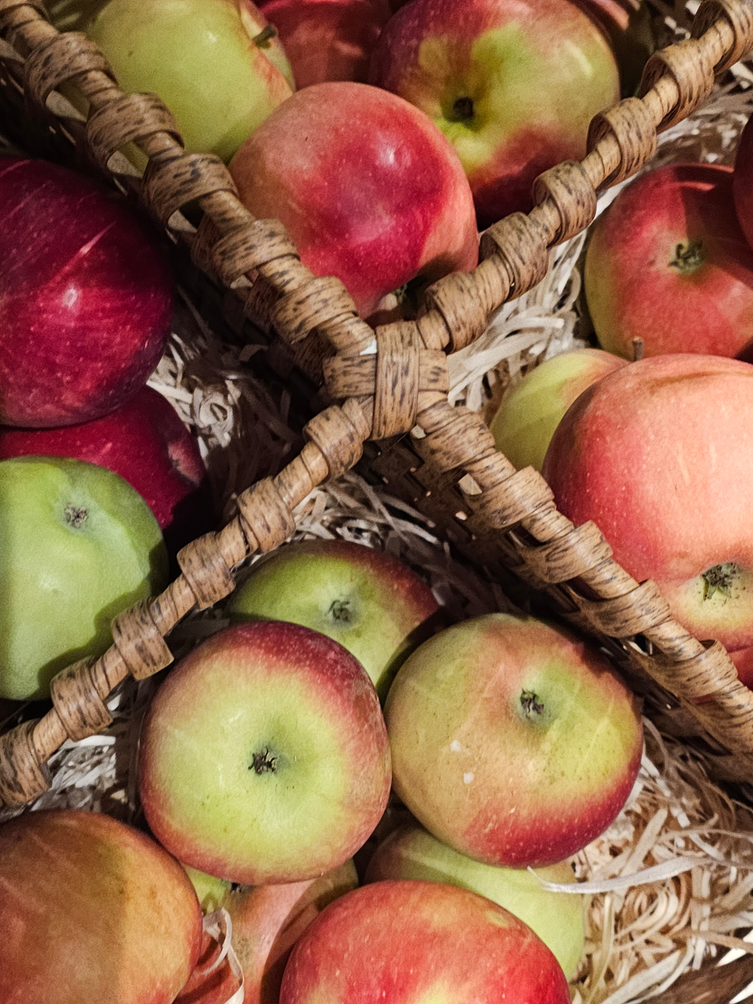 Крупный план свежих красно-зеленых яблок, лежащих в плетеной корзине на соломенной подстилке. Осенний урожай фруктов. Корзина с яблоками с зелено-желтой кожурой и ярким красным румянцем.