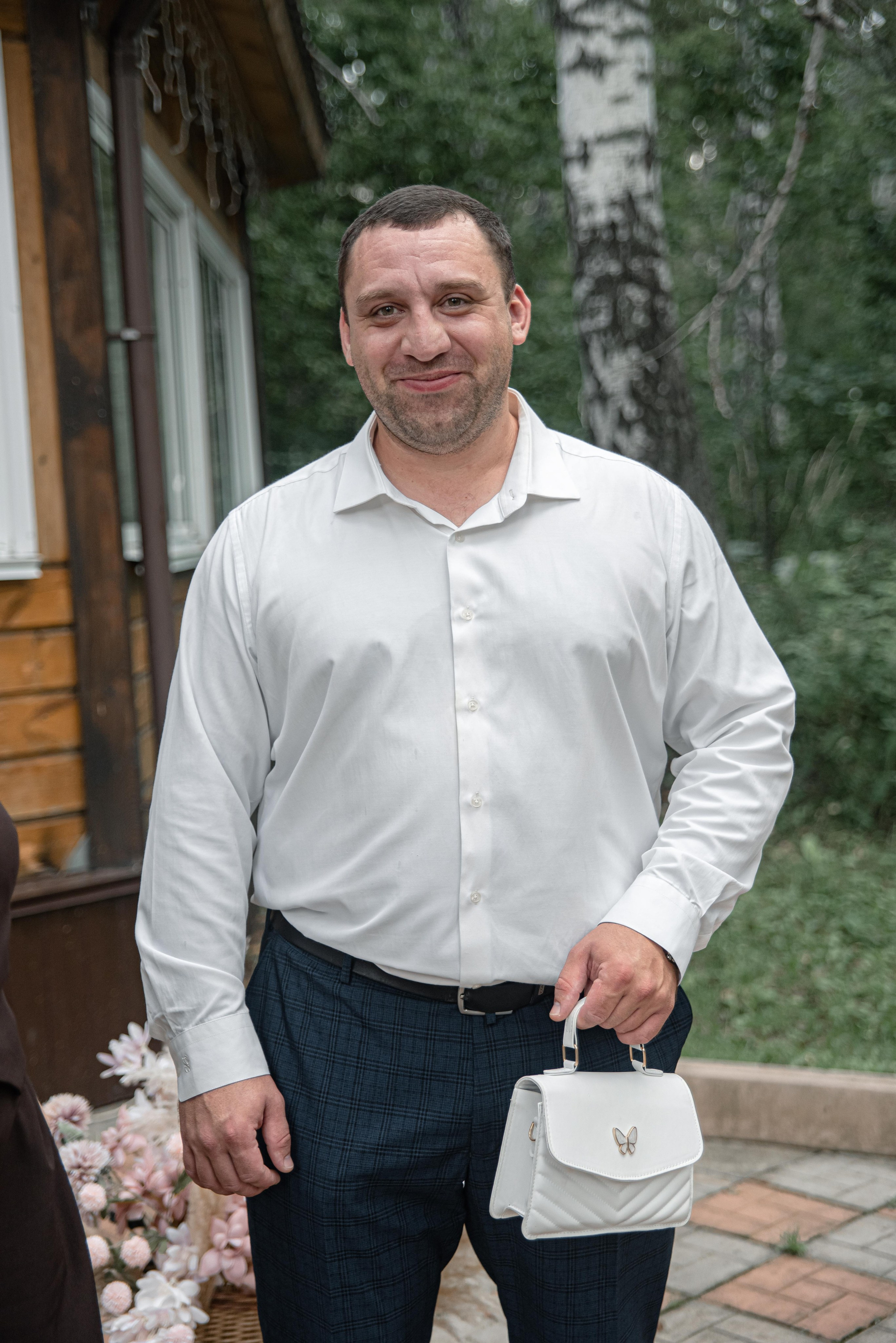 На фото изображен человек в белой рубашке и темных брюках, который держит в руках стильную белую сумочку. Фон состоит из уютного деревянного домика и зелени, создающей атмосферу спокойствия и уюта.