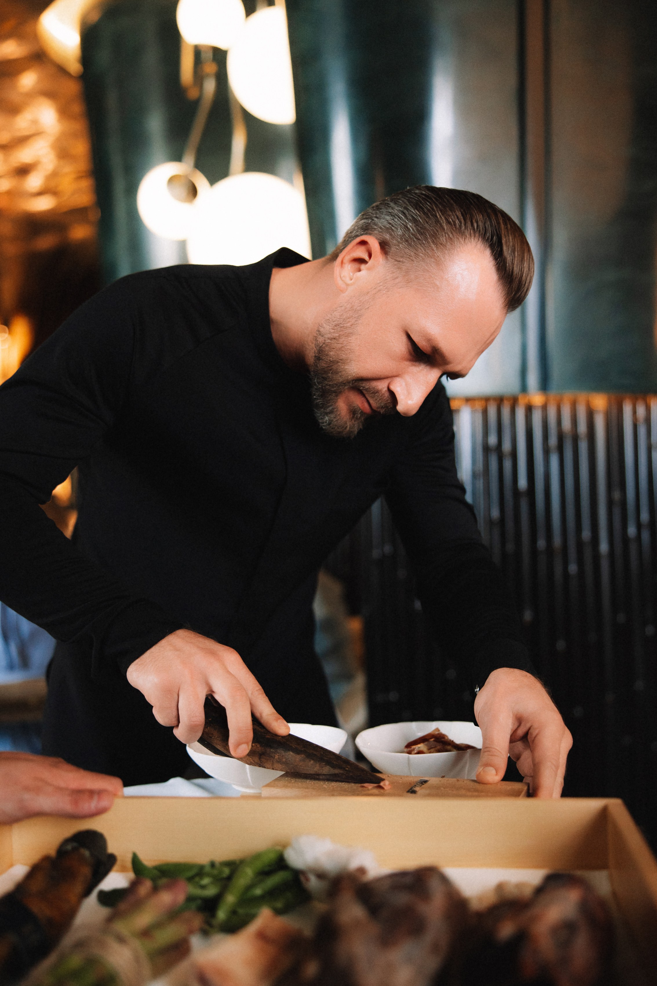 Подача блюда: шеф повар Илья Захаров в черной форме нарезает сушеный тунец для гостей.