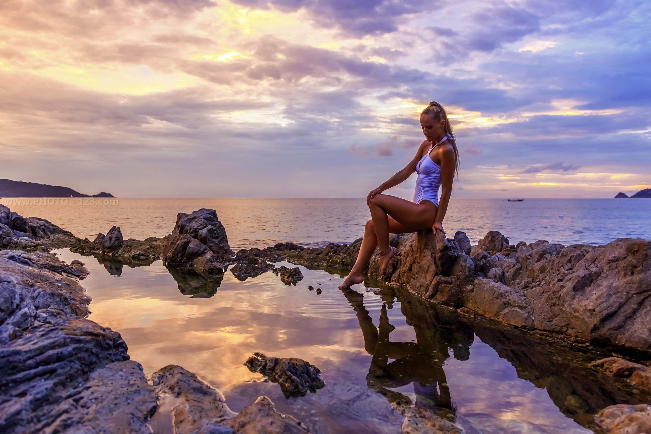 A woman in a light blue one-piece swimsuit sitting on coastal rocks at sunset on Phu Quoc Island, Vietnam. Calm sea and dramatic clouds reflect in a natural tidal pool, creating a serene tropical atmosphere. Outdoor fashion portrait with natural light, pastel sunset tones, and peaceful island scenery.