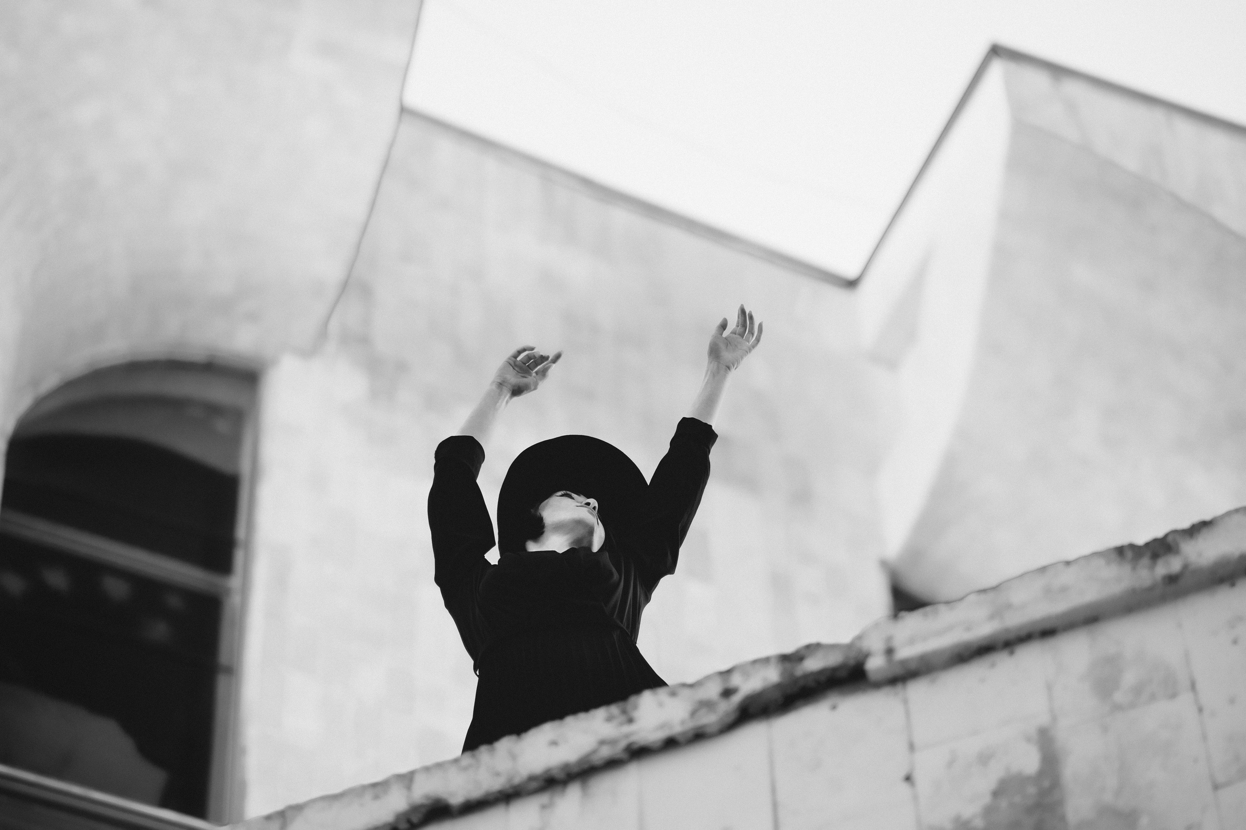 Женщина в чёрной шляпе с поднятыми руками на фоне архитектуры, чёрно-белая концептуальная художественная фотосъёмка