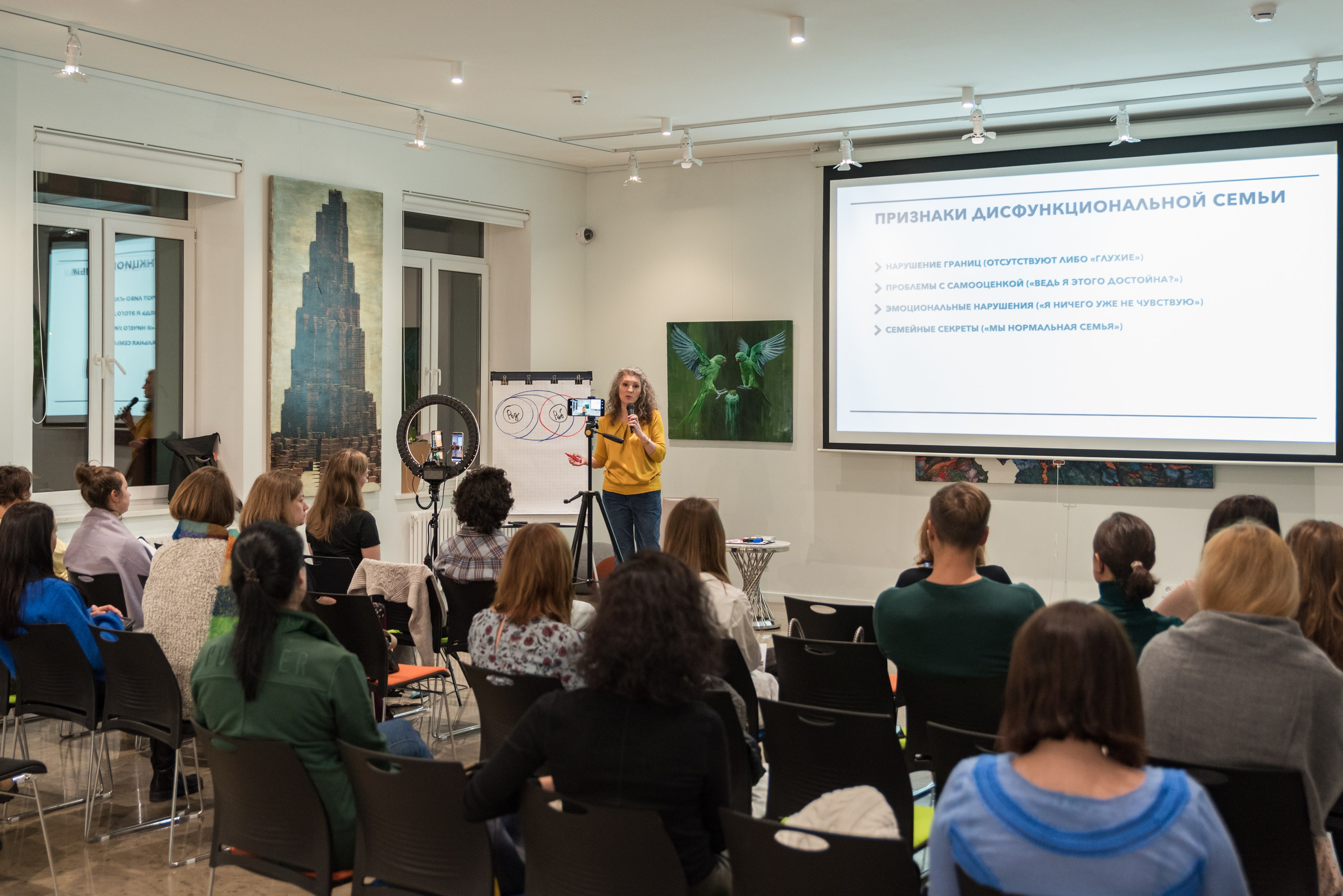 Много зрителей в зале за ними ведущая говорит в микрофон.