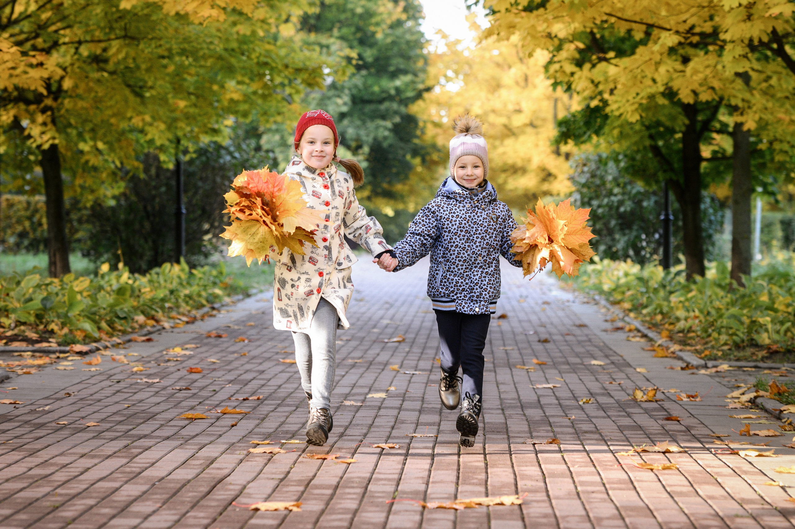 Две девочки бегут по аллее осеннего парка, держа в руках букеты из жёлтых листьев.