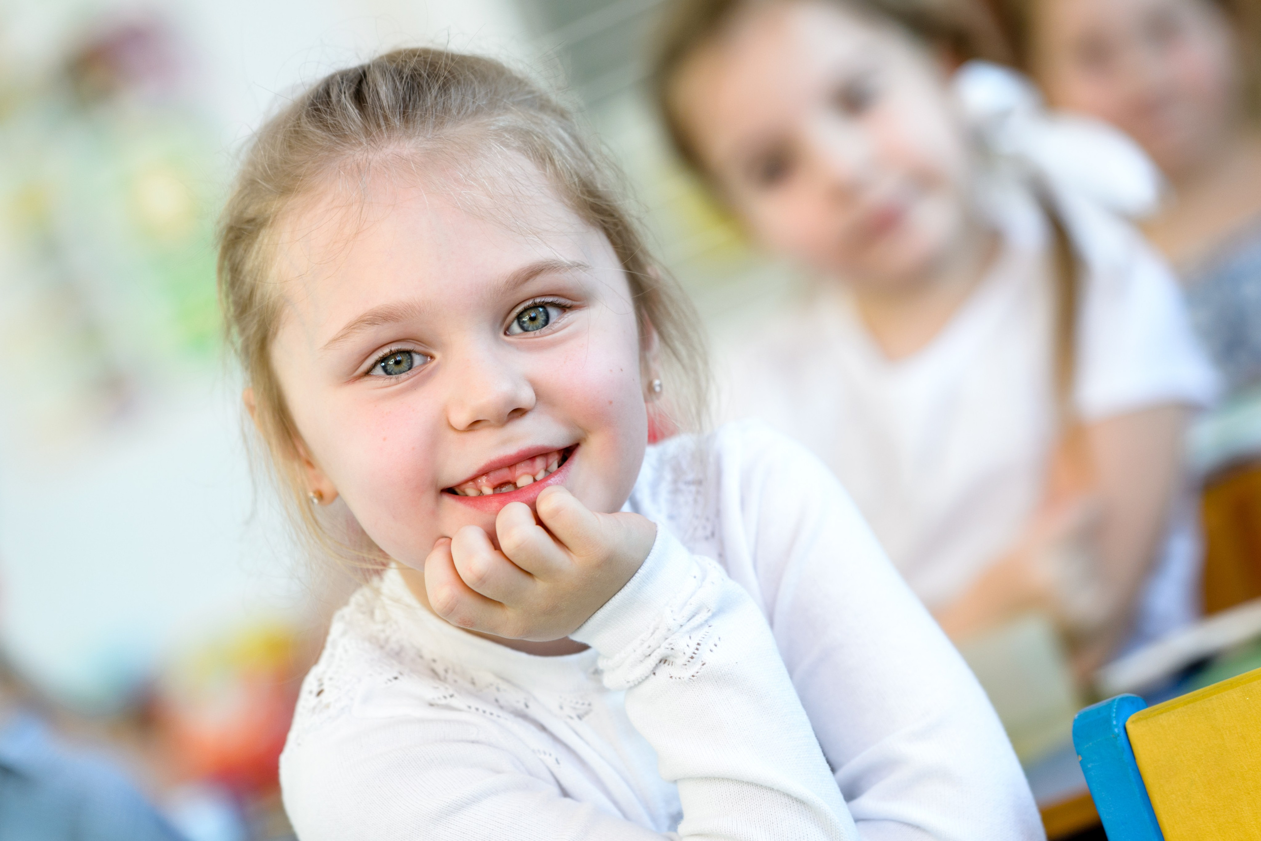 Портрет девочки в группе: улыбается, опираясь на руку, фон с детьми размытым планом.