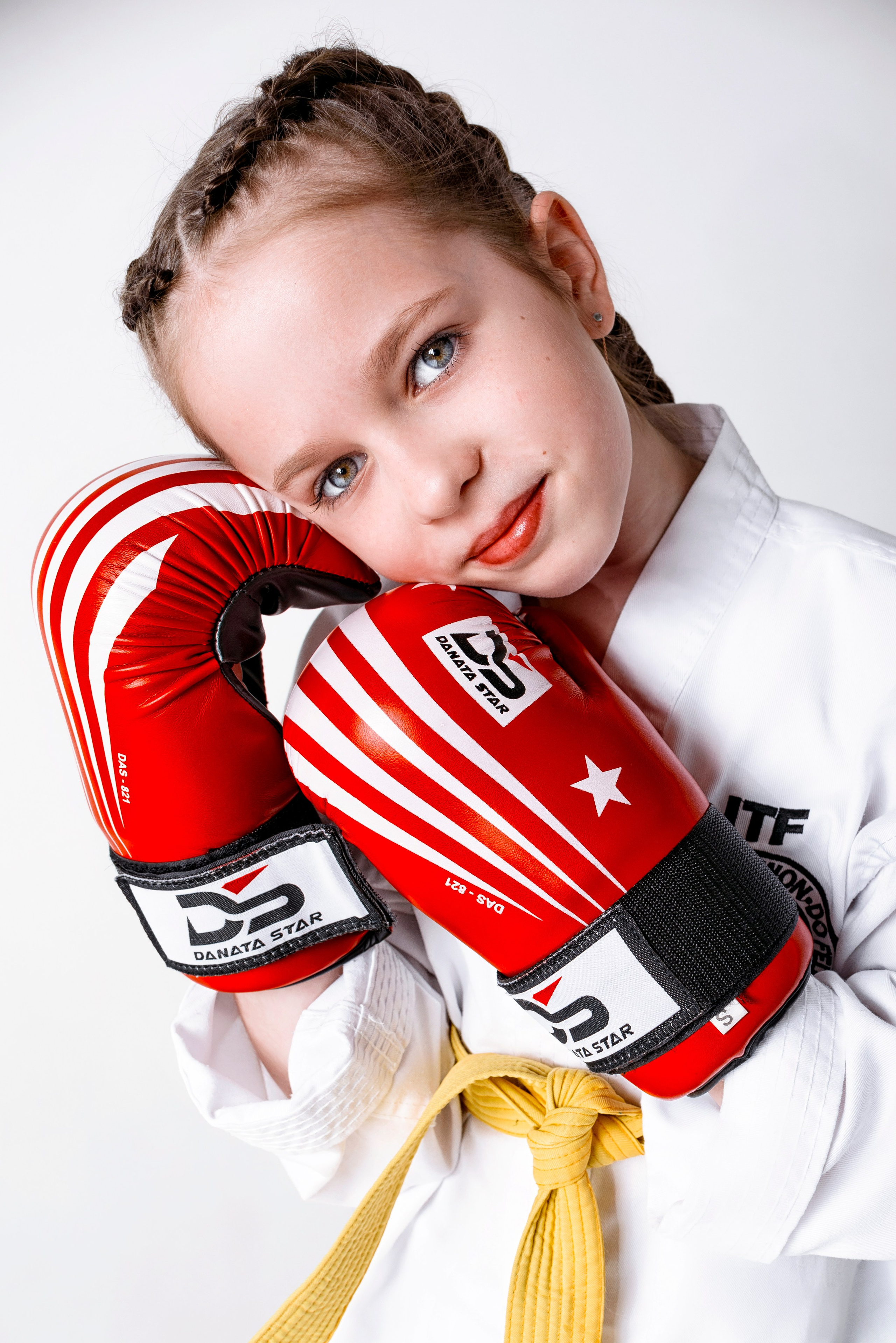 На изображении изображено лицо, одетое в белую форму Тхэквондо (добок) с желтым поясом. Человек носит красные боксерские перчатки и, вероятно, готовится к тренировке боевых искусств. На форме есть нашивки, одна из которых содержит буквы "ITF", что, вероятно, указывает на Международную федерацию Тхэквондо. Фон светлый и однородный, что обеспечивает четкий обзор объекта.