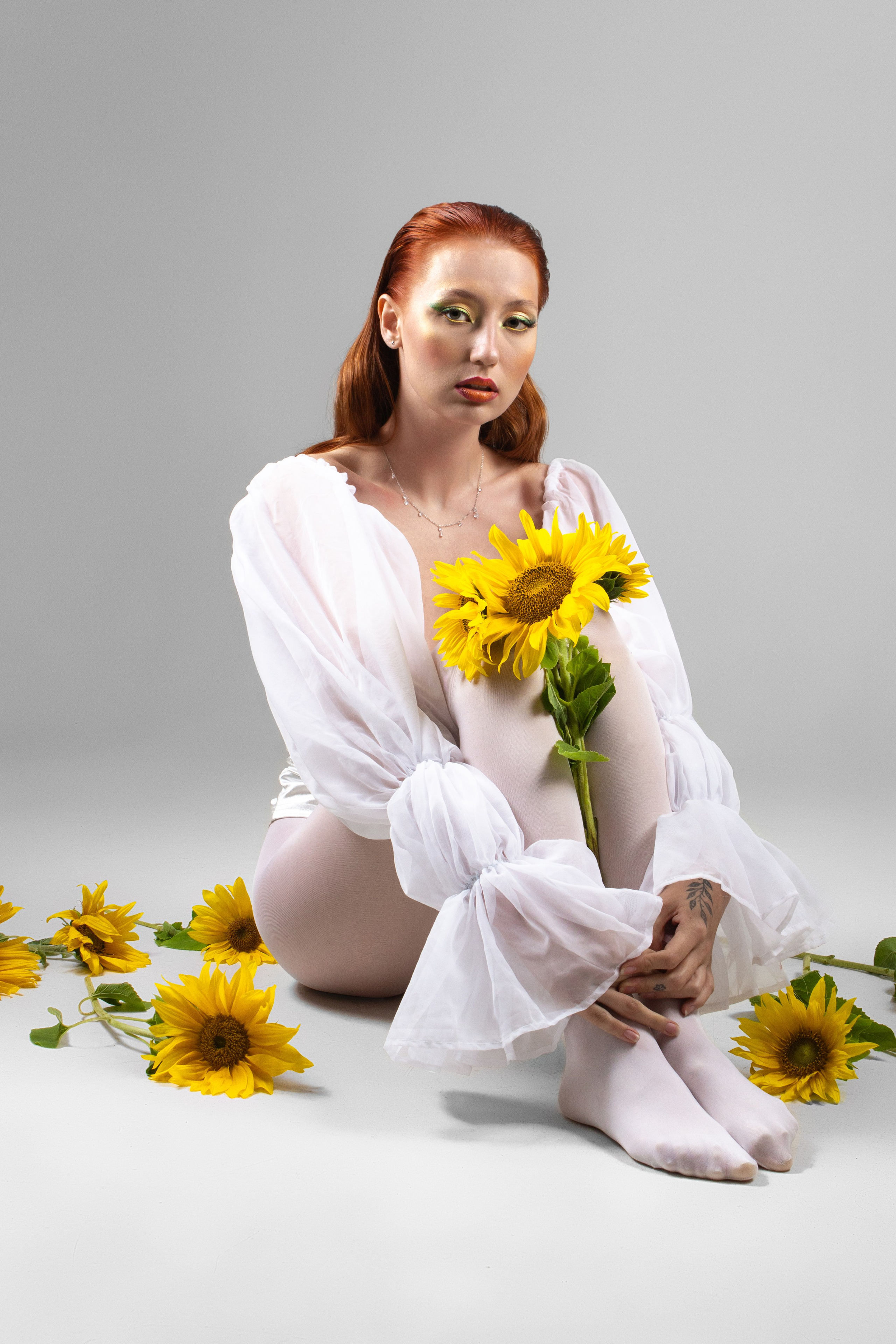 стройная девушка в белой одежде обнимает себя за колени, возле нее лежат несколько подсолнухов 