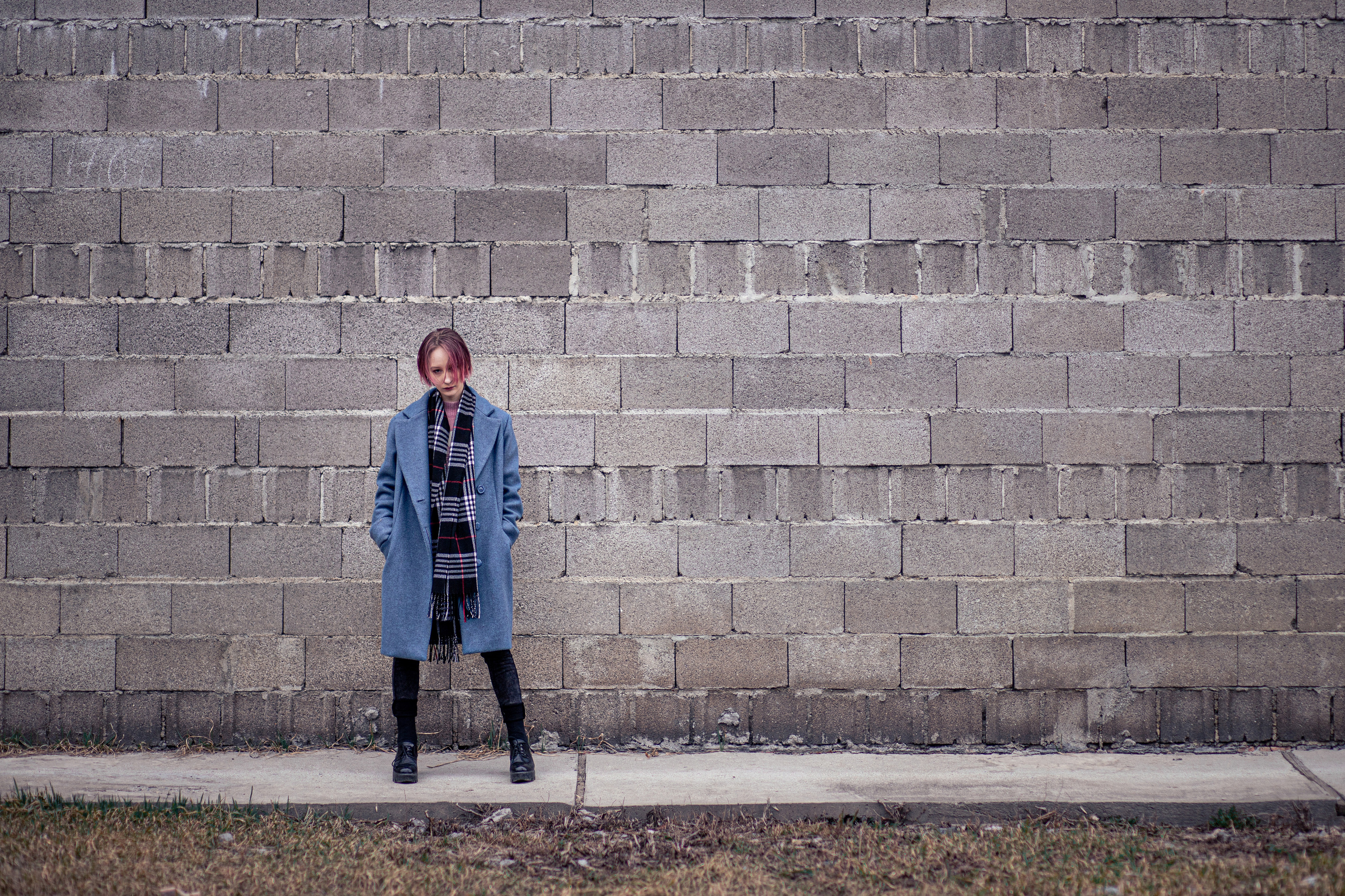 Девушка с розовыми волосами в синем пальто на фоне серой стены из пеноблока томно смотрит в кадр