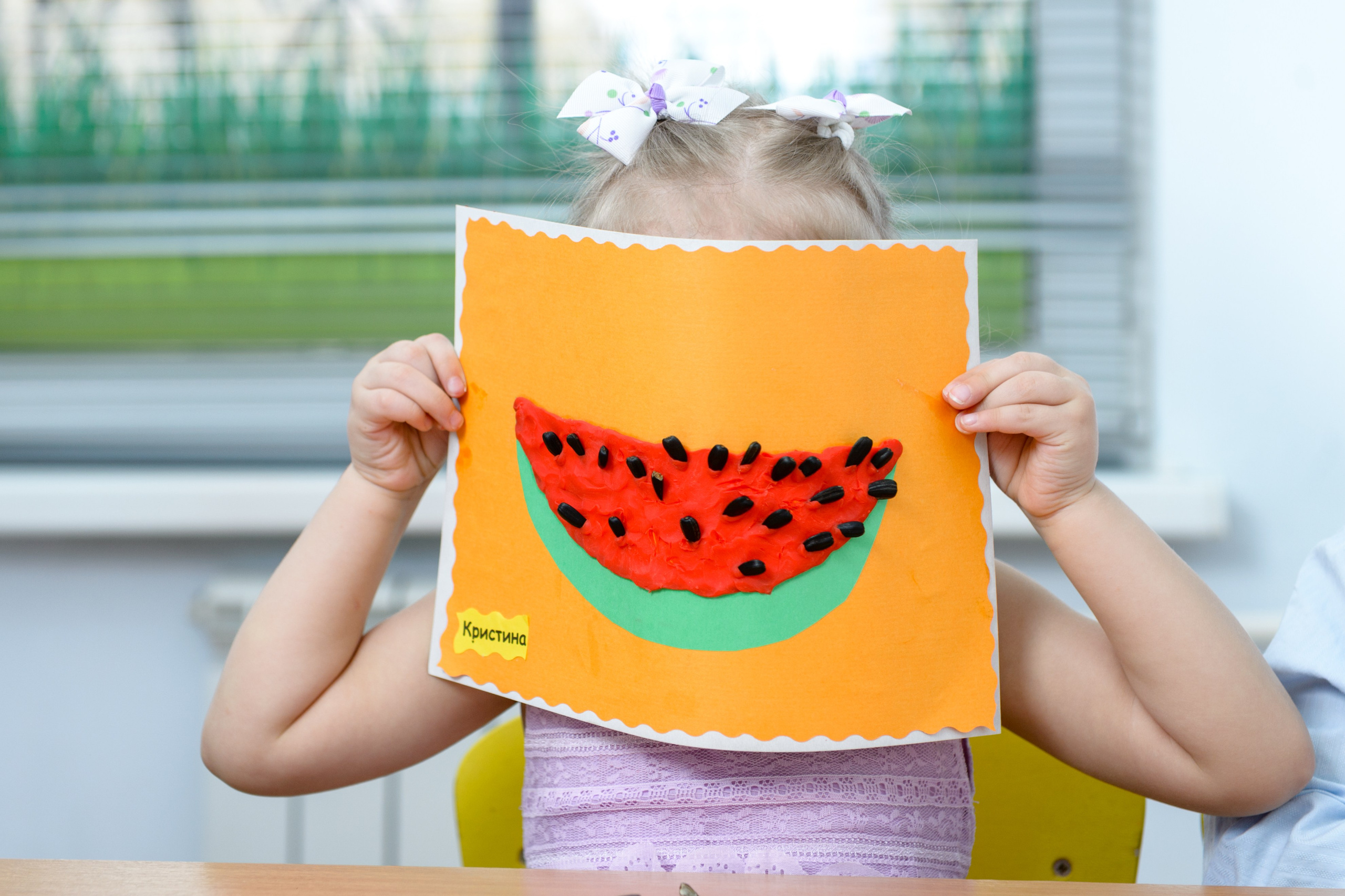 Девочка держит поделку из бумаги в виде арбузика, закрывая ею лицо на фоне окна. 