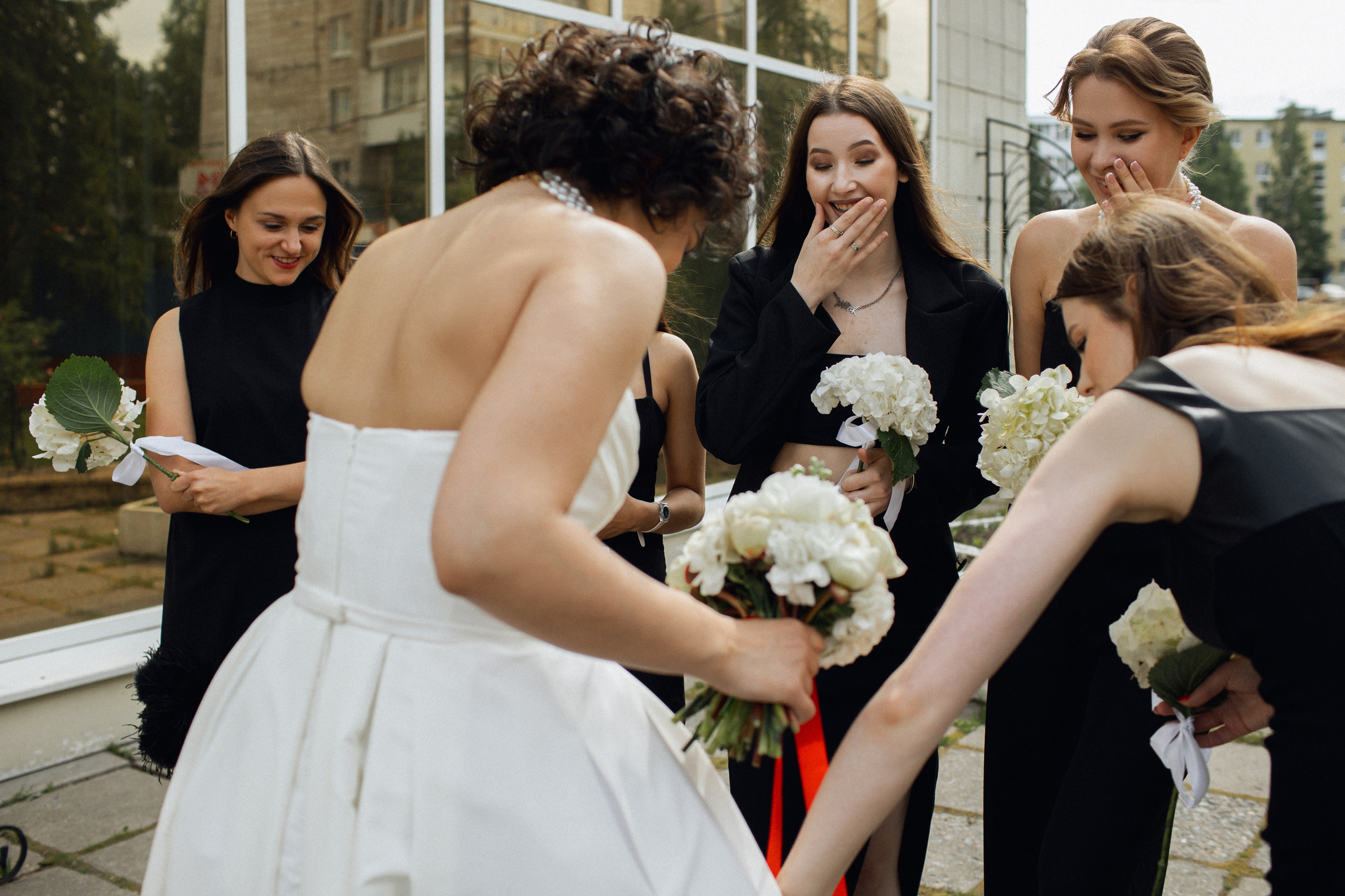 Свадебный бьюти-момент: подружки в черных платьях рассматривают атласное платье невесты от салона NATALIE L 