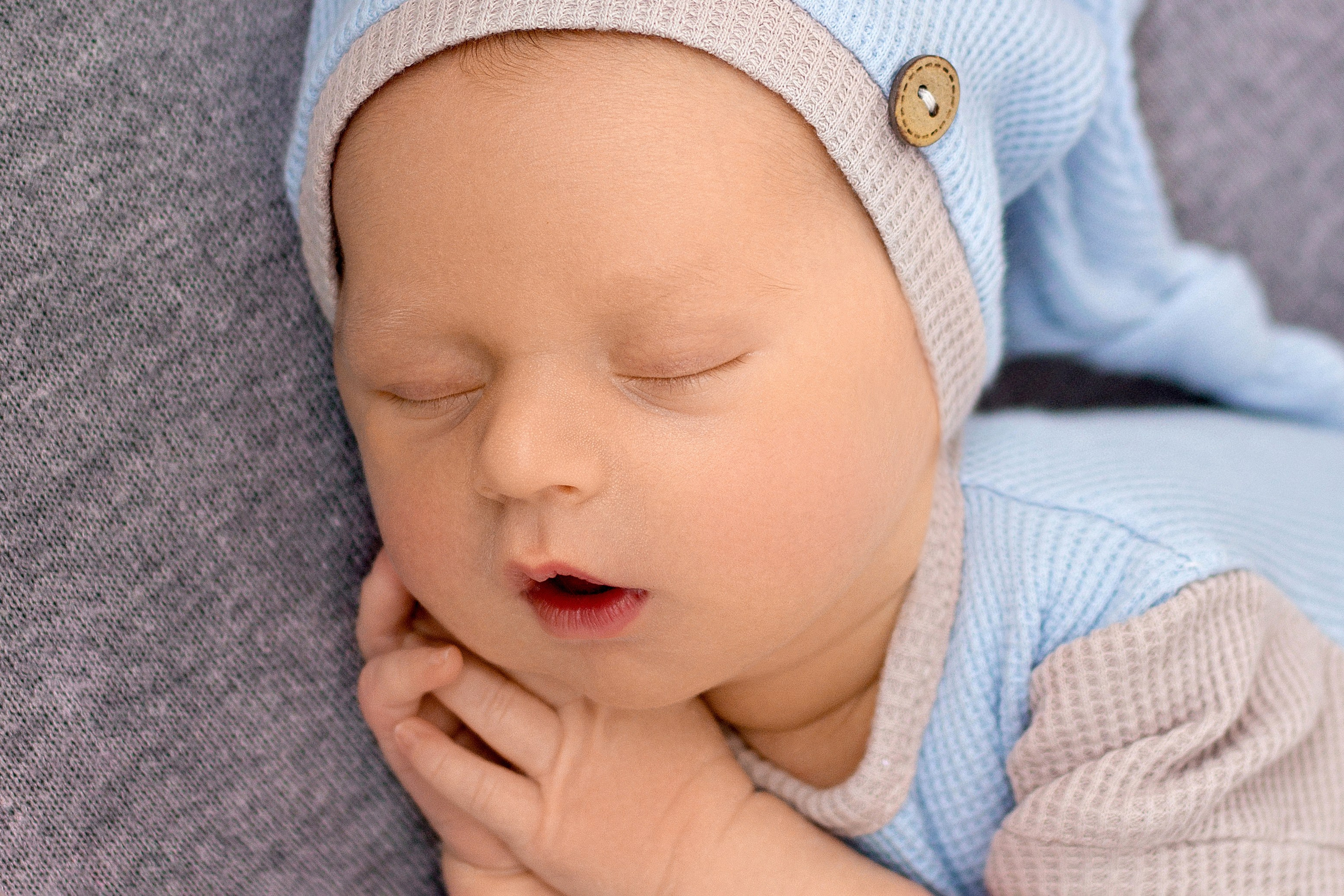 портрет новорожденного с коликами в голубо-сером костюмчике, после успокоения, с ручками под щечкой на сером фоне и с колпачком на голове