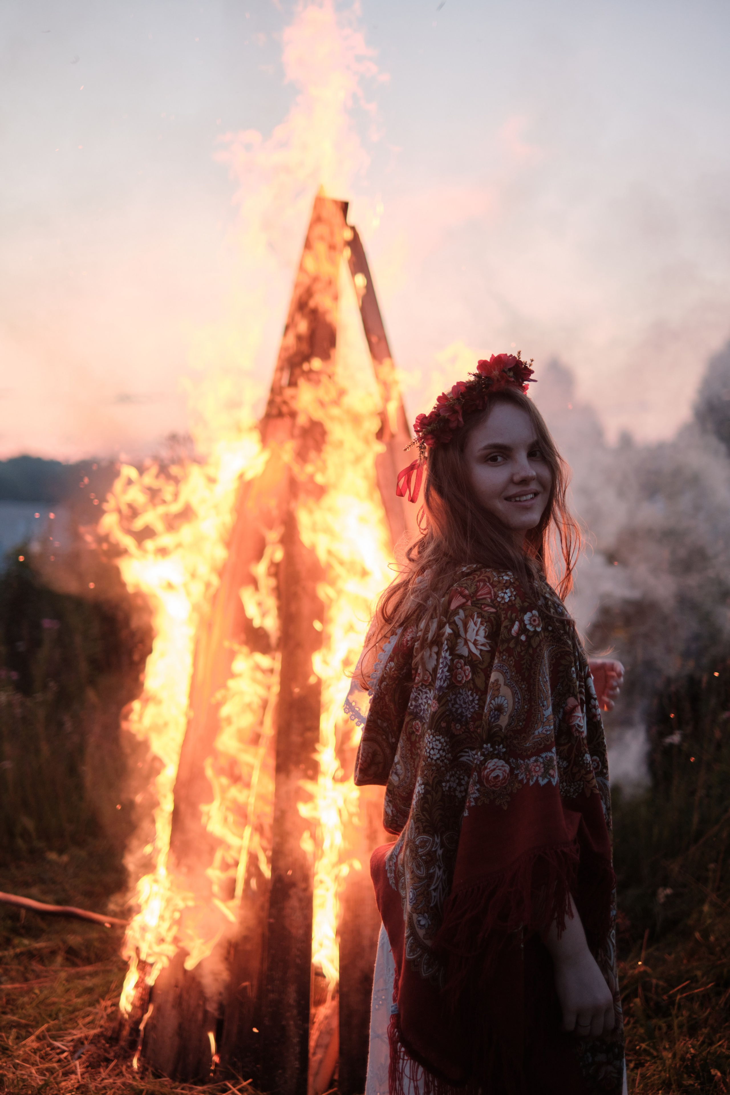 Девушка стоит в русском костюме и венком сзади горит большой костер