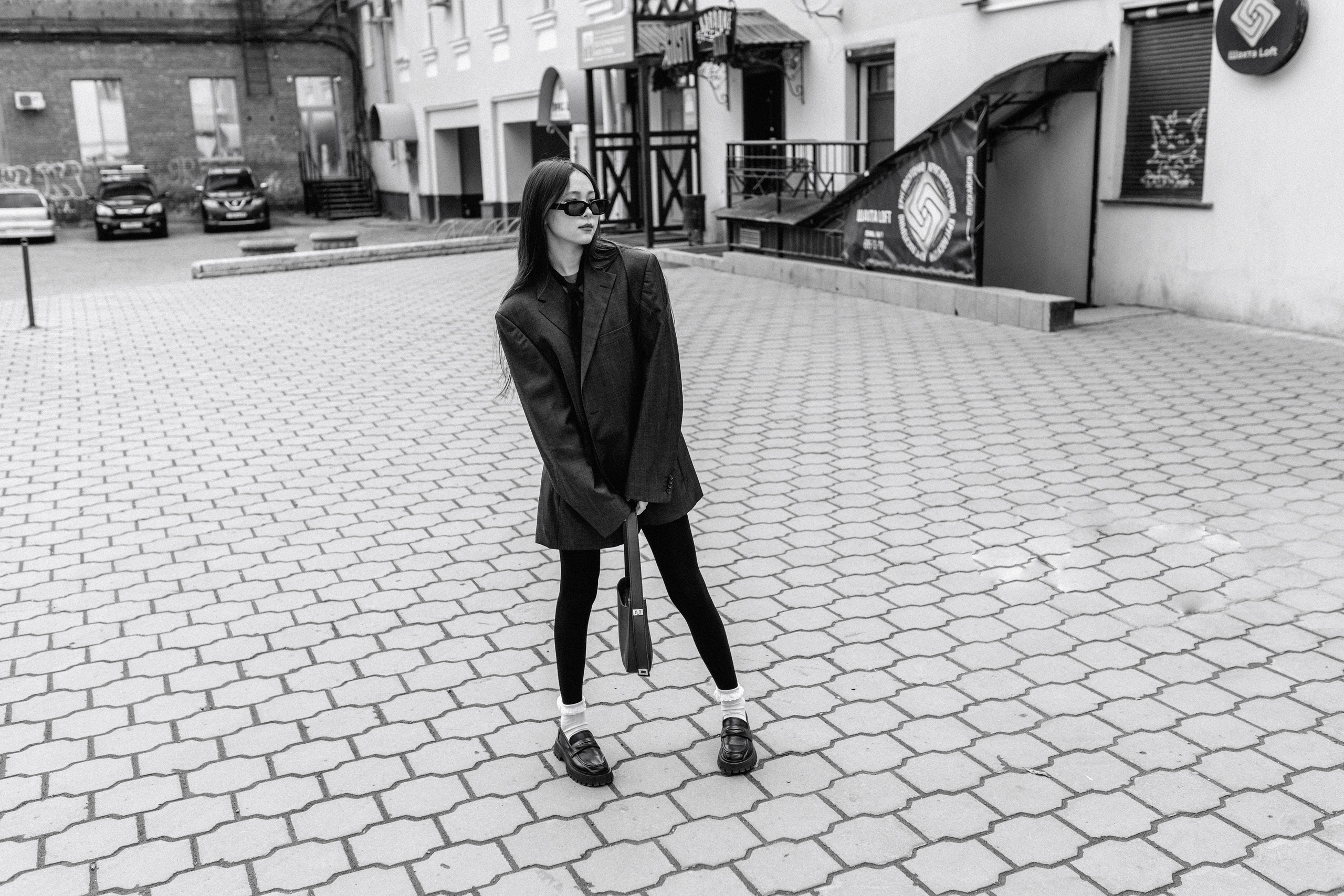Черно-белое изображение  девушки в блейзере оверсайз, леггинсах и лоферах, уверенно позирующей  в городском дворике. Настроение стильное и острое.