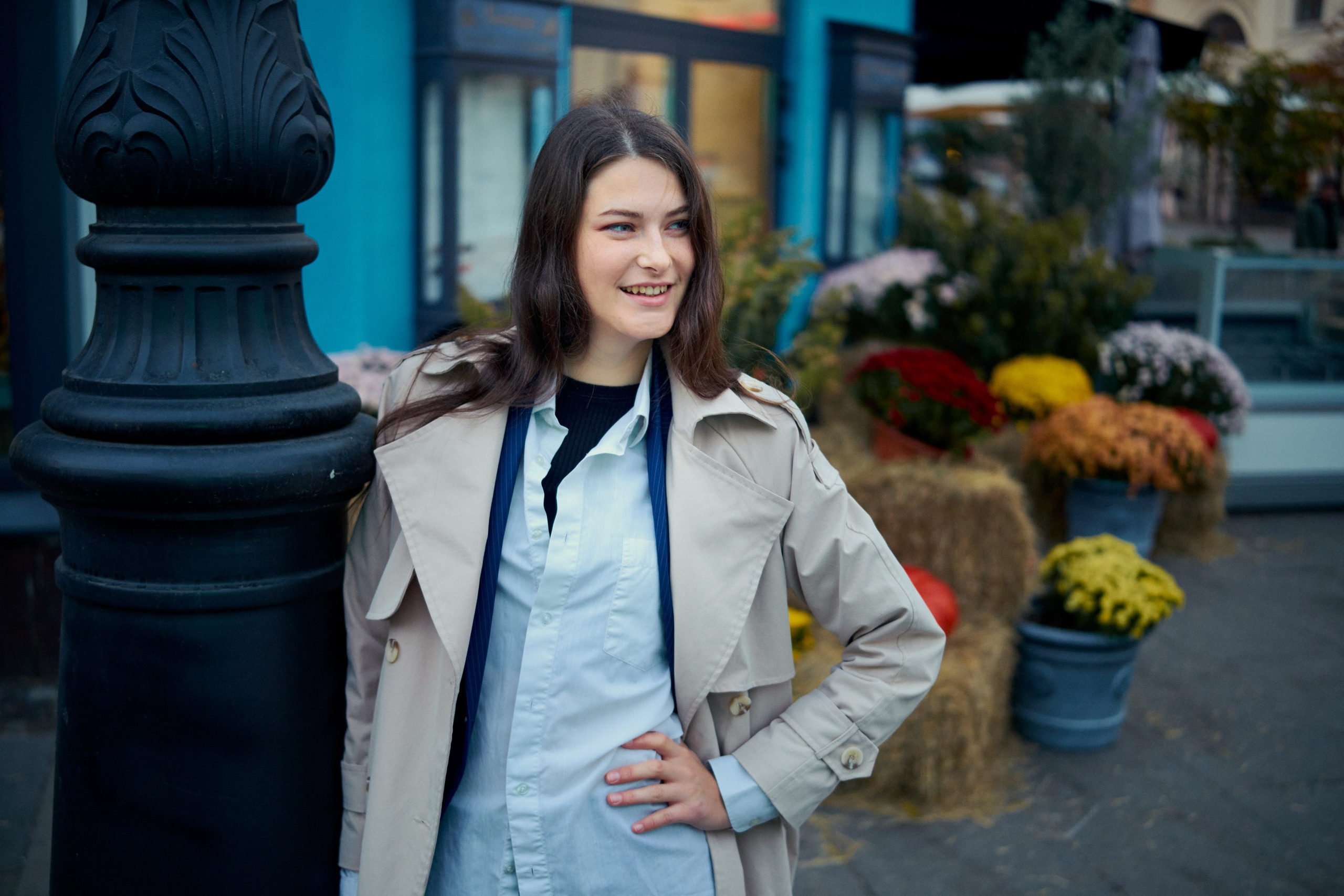  Элегантная девушка в светлой рубашке и пальто, позирует рядом с фонарным столбом в городской среде