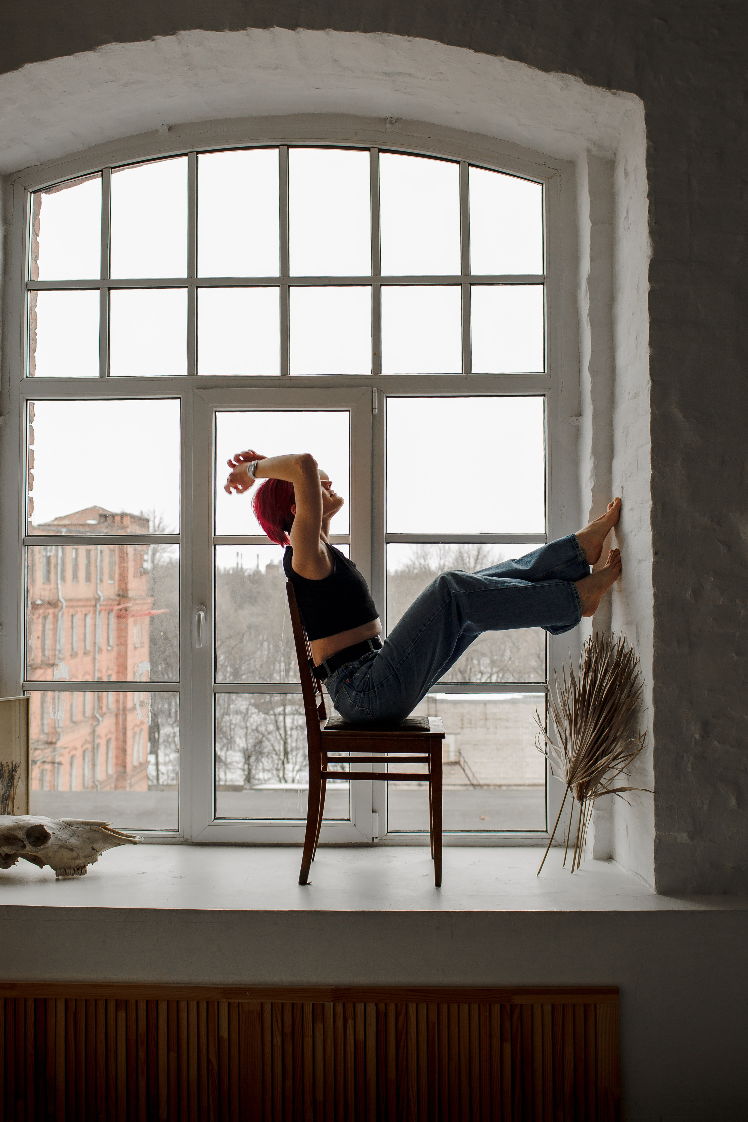 Портрет девушки в полный рост; она сидит у панорамного окна, приняла необычную позу.