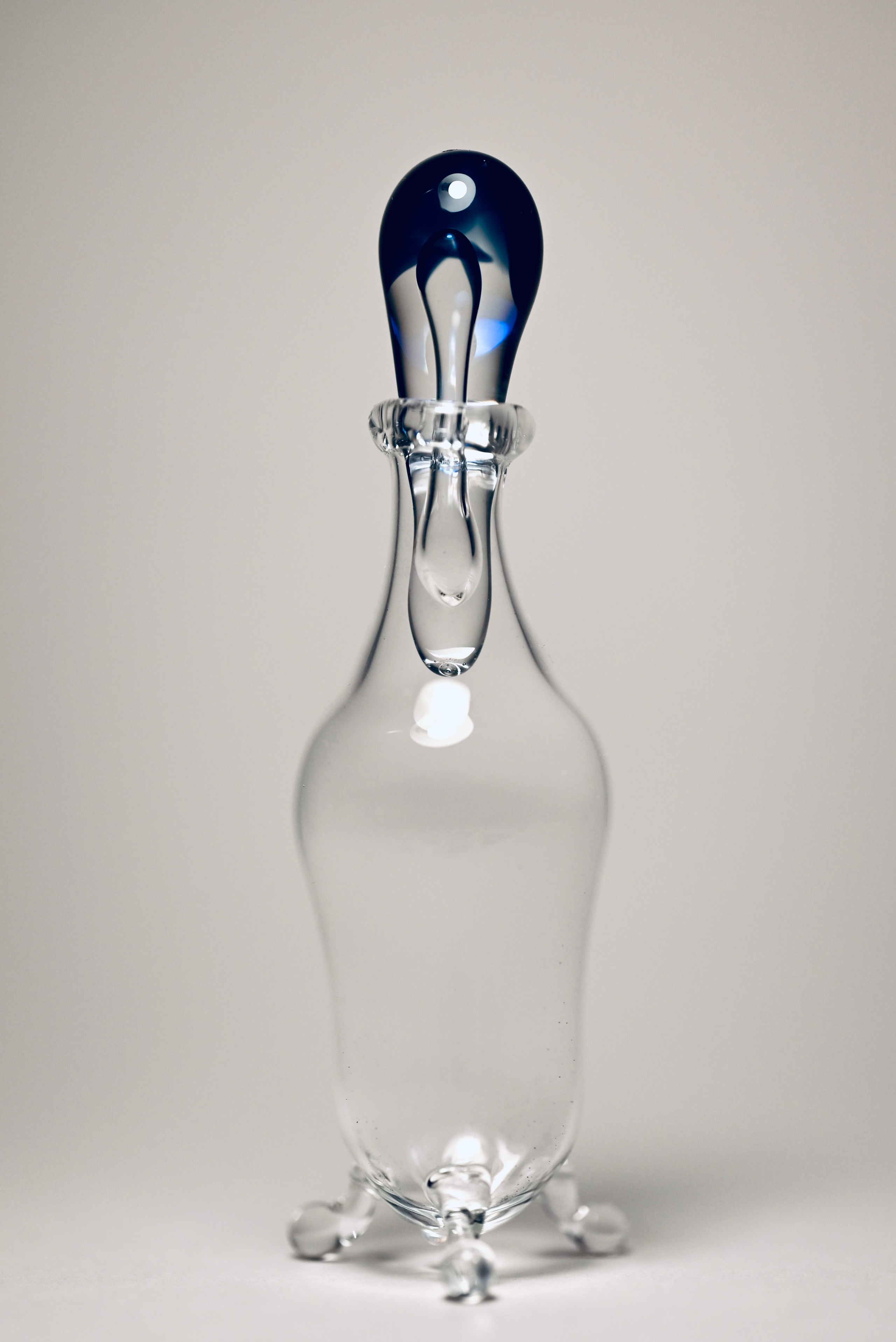Флакон «Ниоба». Боросиликатное стекло. 2025 г. Флакон из прозрачного боросиликатного стекла с каплевидной пробкой насыщенного синего цвета. Форма напоминает слезу.Название отсылает к древнегреческому мифу о Ниобе. Объект может использоваться как декоративный сосуд, а также для хранения эссенций, ароматических масел или других жидкостей.Данный флакон визуально ассоциируется с темой чистоты, тишины и хрупкости.