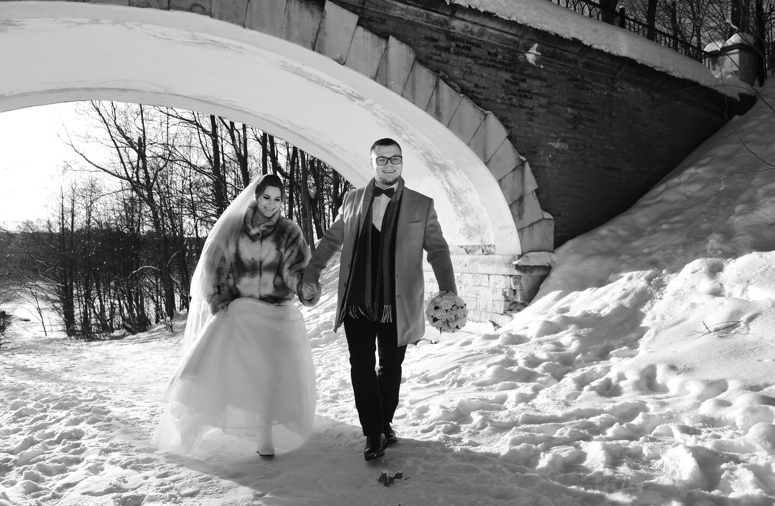 Зимняя свадьба в усадьбе Суханово в лютый мороз, жених и невеста идут по зимней дороге и держатся за руки на фоне арки моста, черно-белая фотография