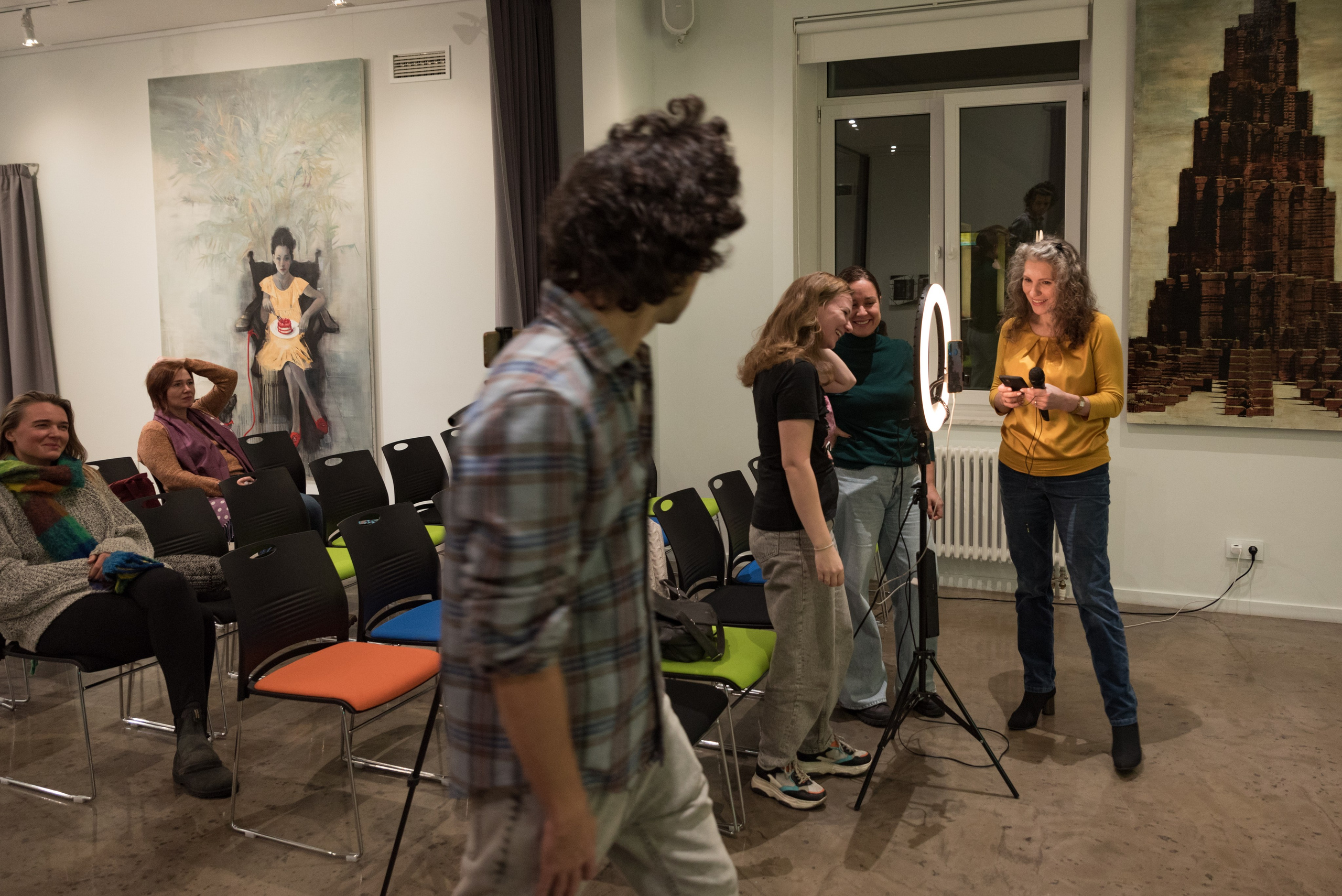 Подготовка к началу семинара по психологии. Мужчина идёт на переднем плане. На заднем стоит ведущая с микрофоном и помощники настраивают освещение.