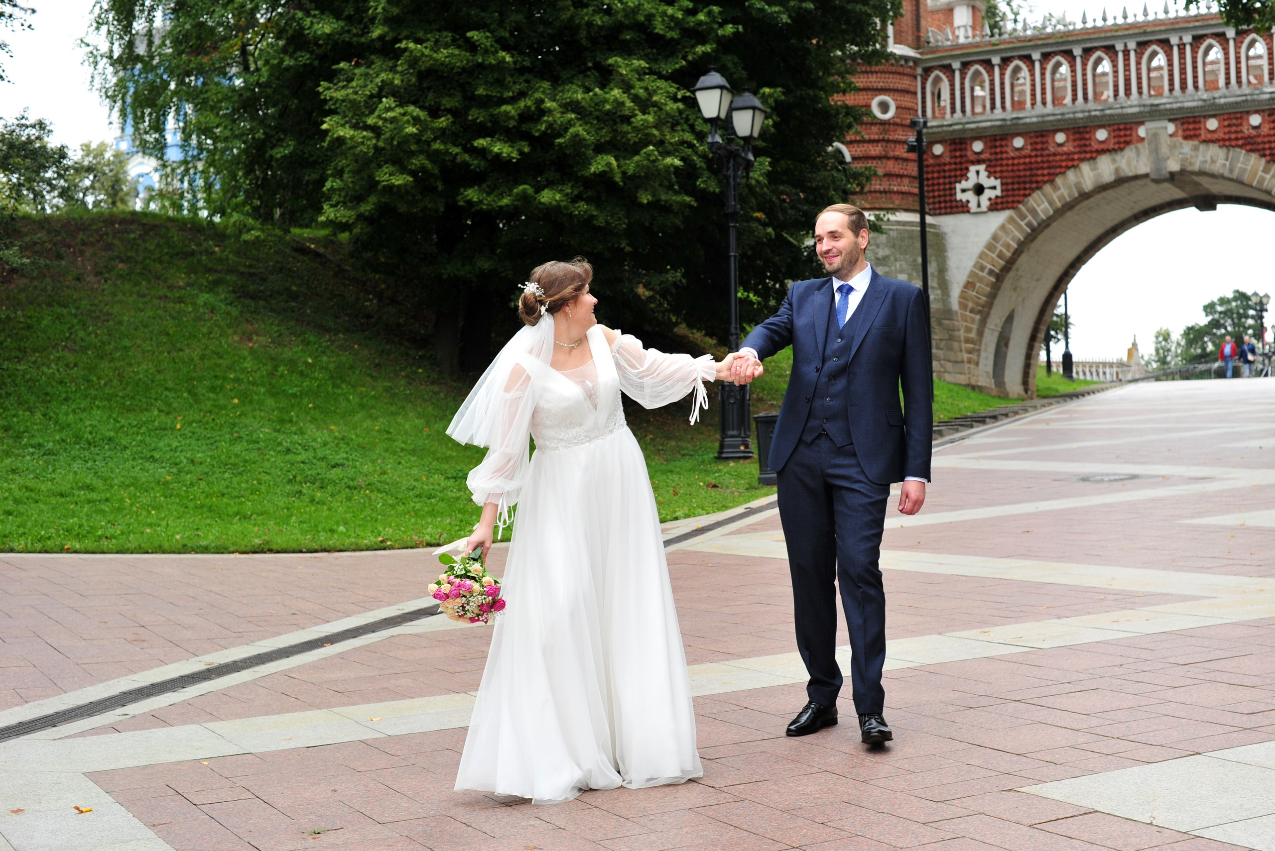 Свадьба в Царицыно пасмурным летним днем, невеста идет впереди жениха и тянет его за руку за собой на фоне моста из красного кирпича 