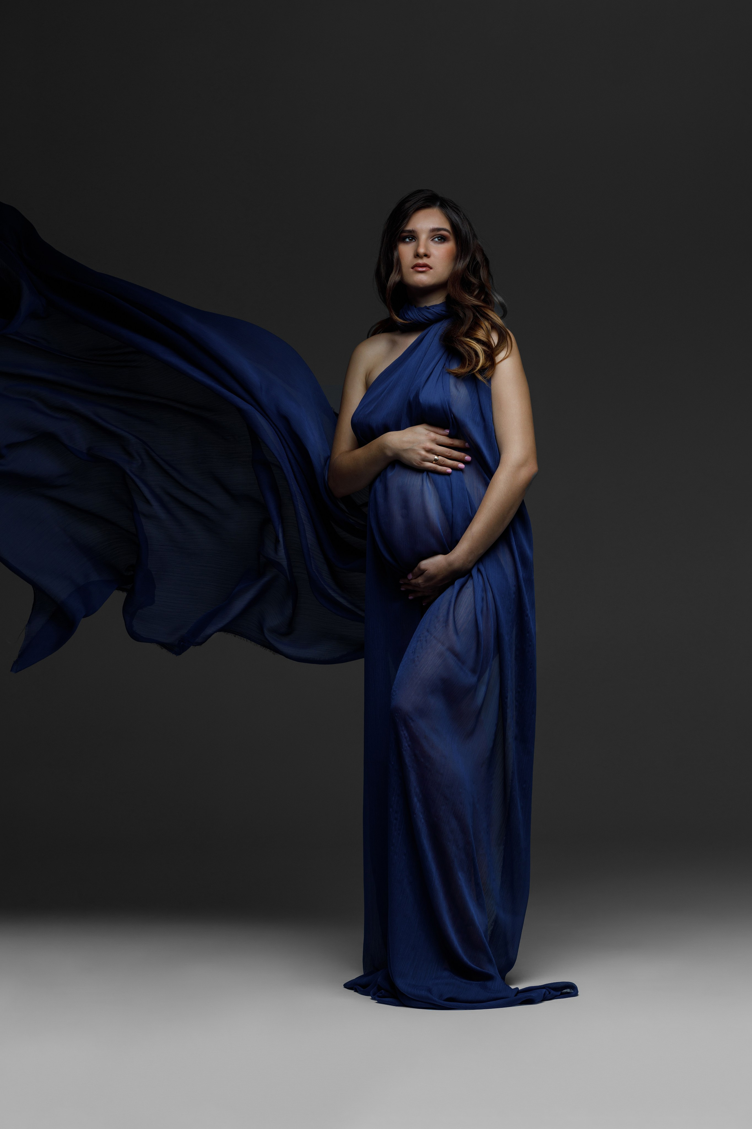 Беременная девушка стоит в тканях на темном фоне и держит животик