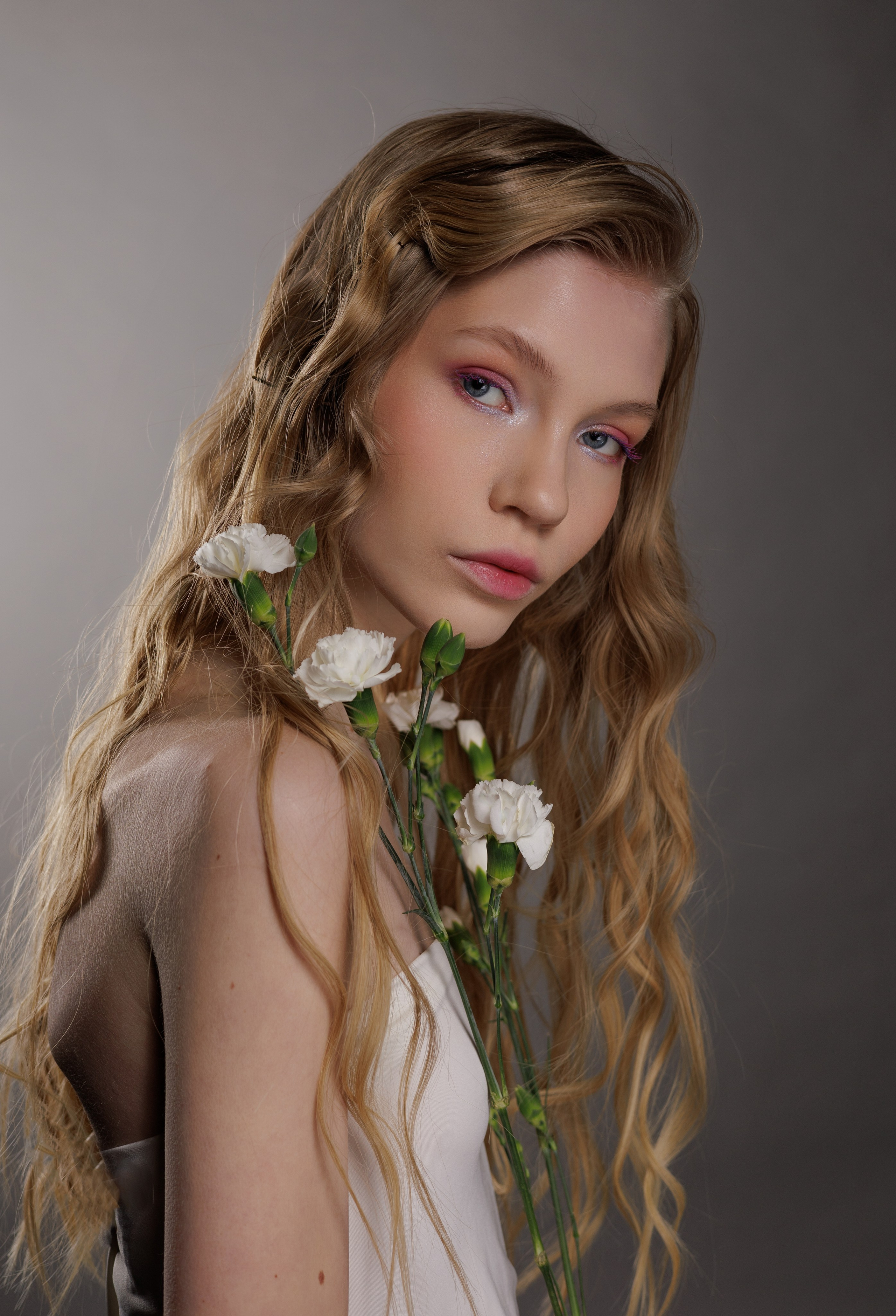 Молодая женщина с длинными волнистыми светлыми волосами и нежно-розовыми тенями на веках мягко отводит взгляд в сторону. Белые цветы свисают с ее плеча, создавая элегантное, безмятежное настроение.