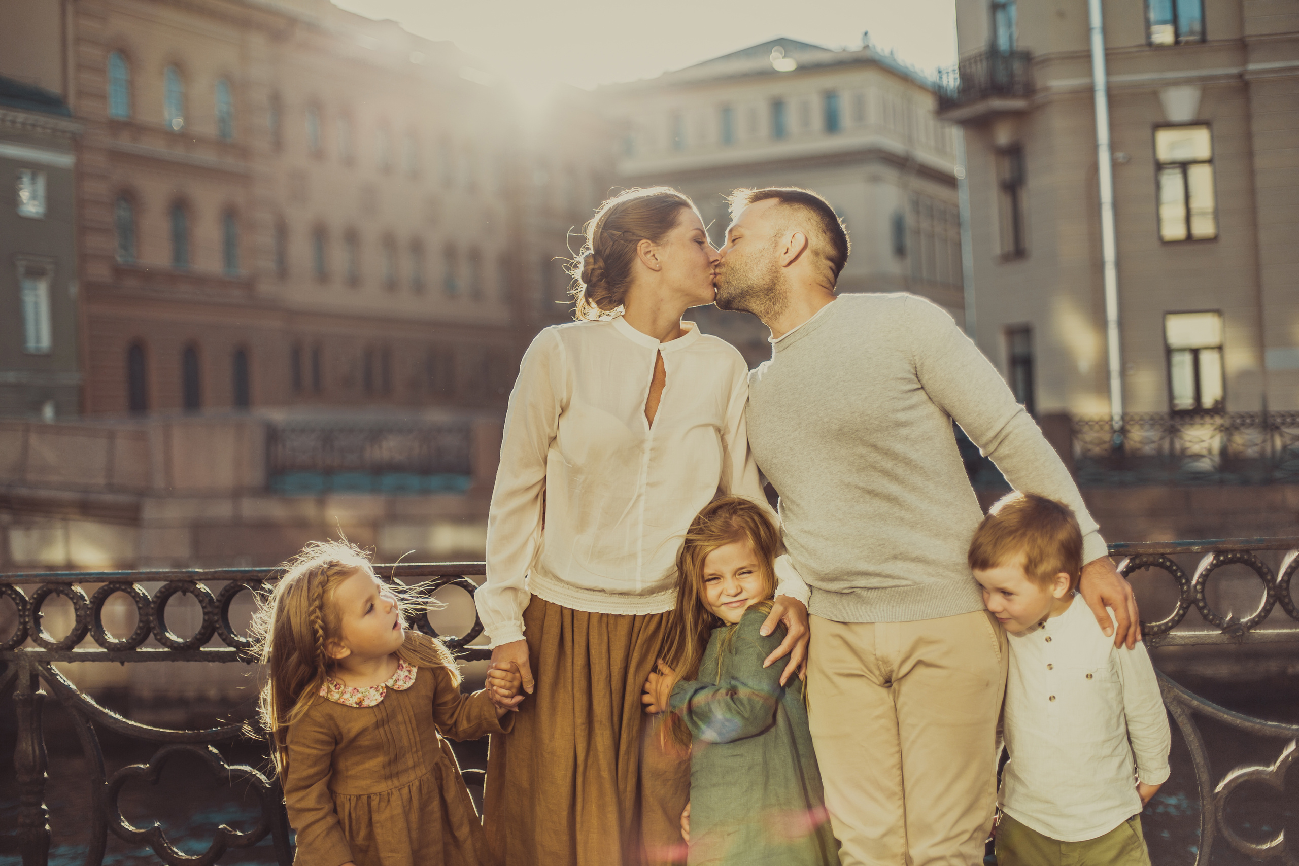 родители стоят на набережной Санкт-Петербурга со своими маленькими детьми в солнечный день и целуются