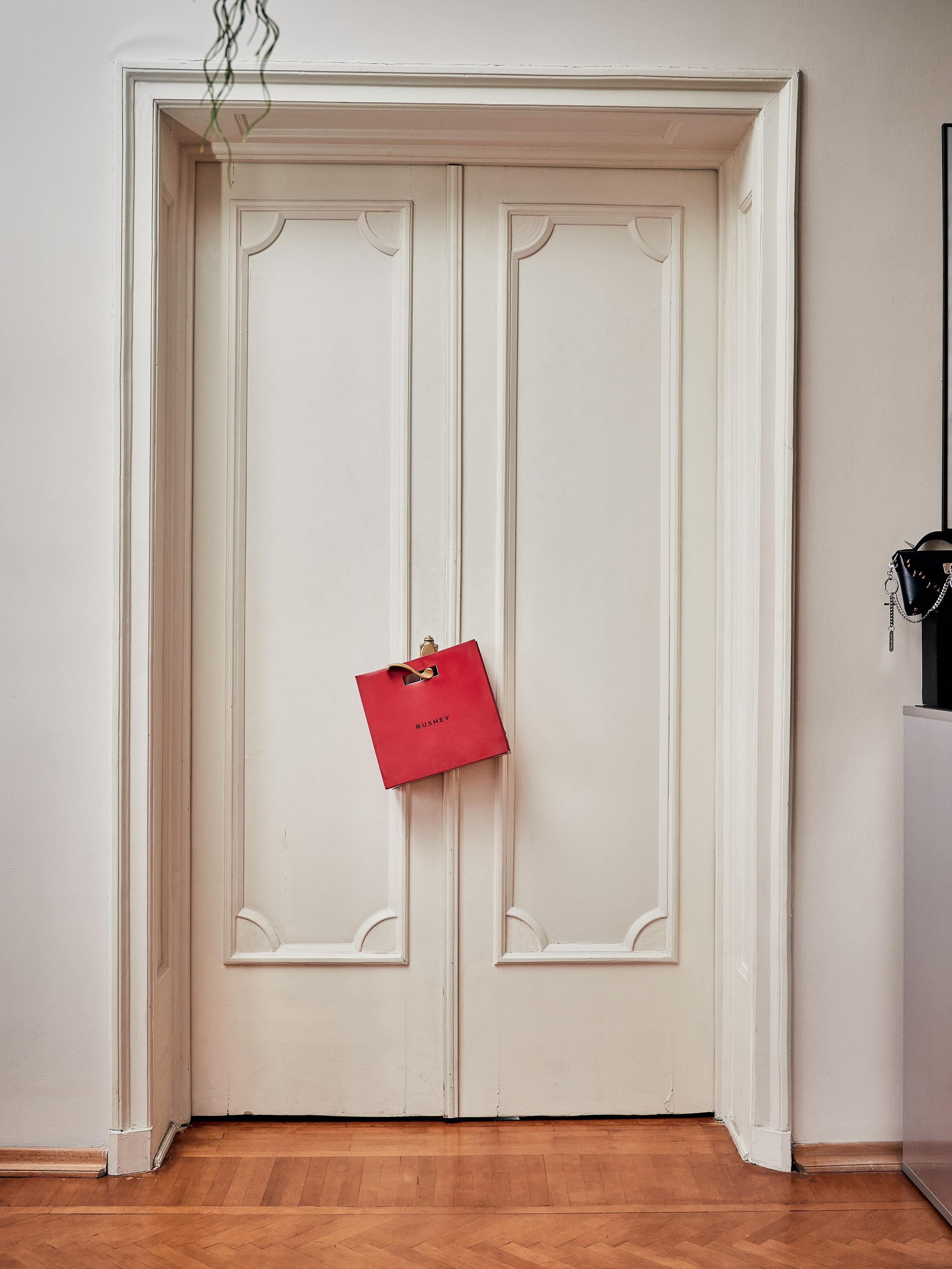 Пара белых двойных дверей с замысловатыми панелями, закрытых и обрамленных белой стеной и плинтусом. На ручке висит красная сумка для покупок с надписью «Givenchy». Пол имеет полированную деревянную отделку. На правом краю виден частичный проблеск черного объекта.