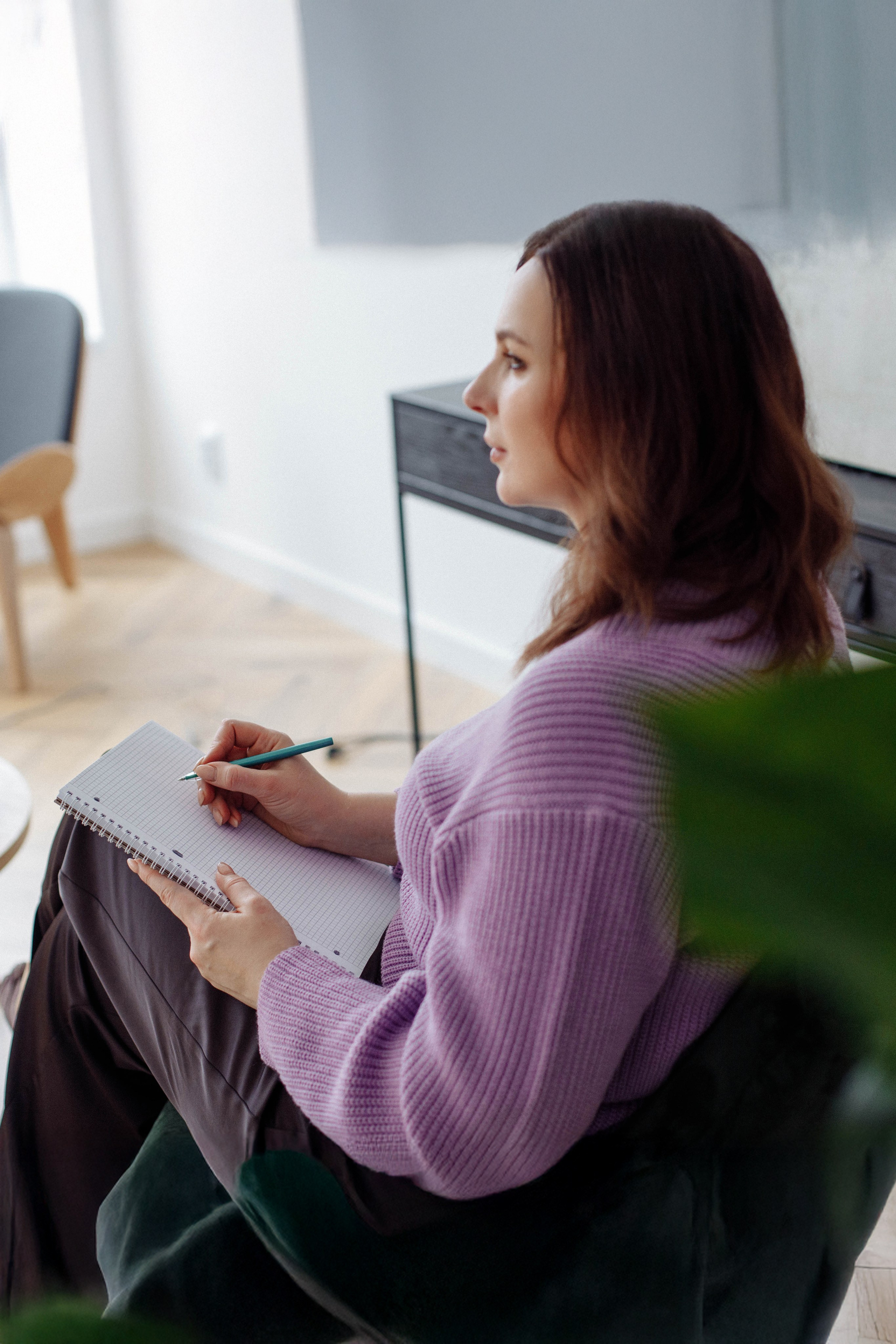 Задумчивый образ: психолог с блокнотом в руке, сидя боком, созерцает вид из окна студии.