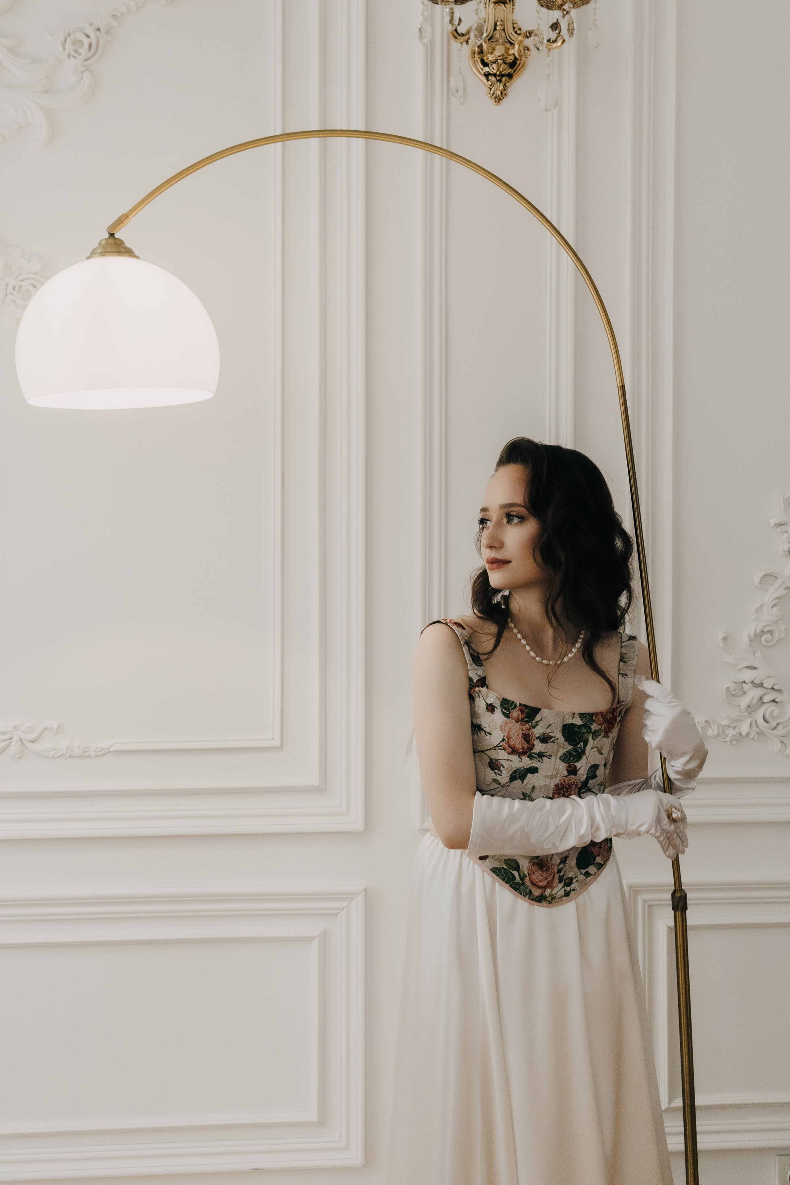 Фотограф Александра Овечкина Пенза: модель стоит рядом с изящным золотым торшером в светлой классической комнате.