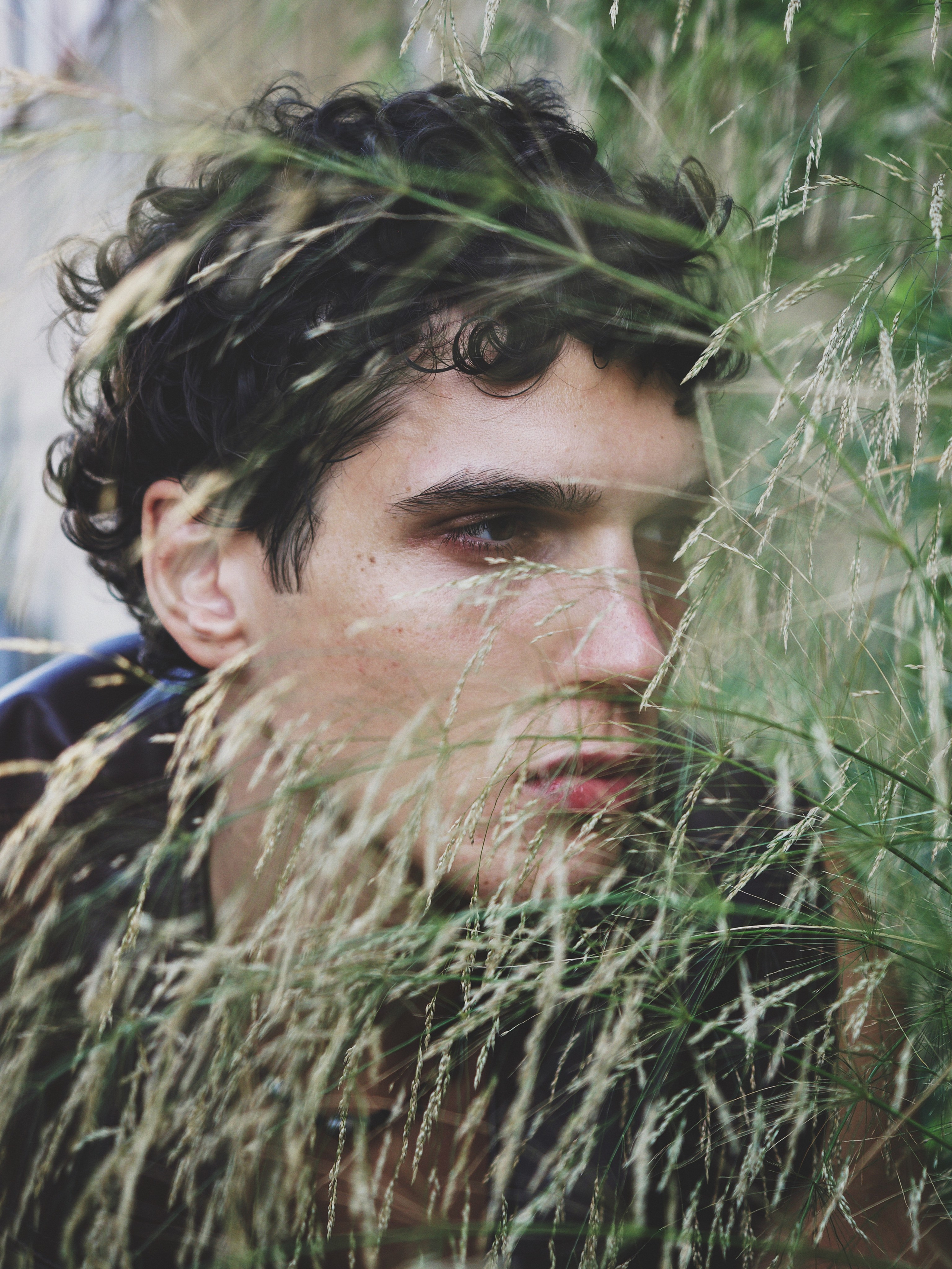 Close-up portrait of Cem Kurttekin surrounded by grass for a fashion editorial