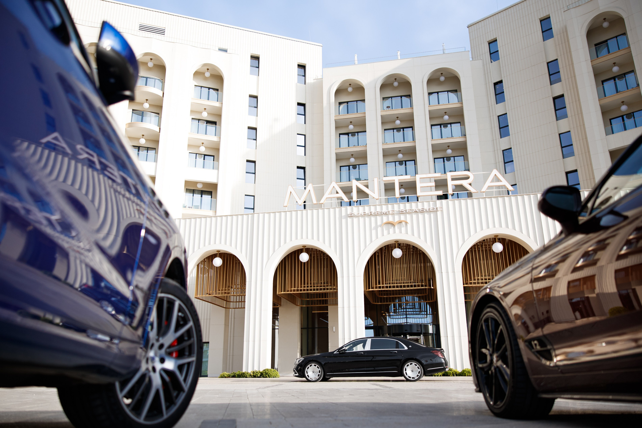 Экстерьер: Вид на фасад отеля Mantera Supreme через колеса элитных автомобилей, подчеркивающий премиальность локации.