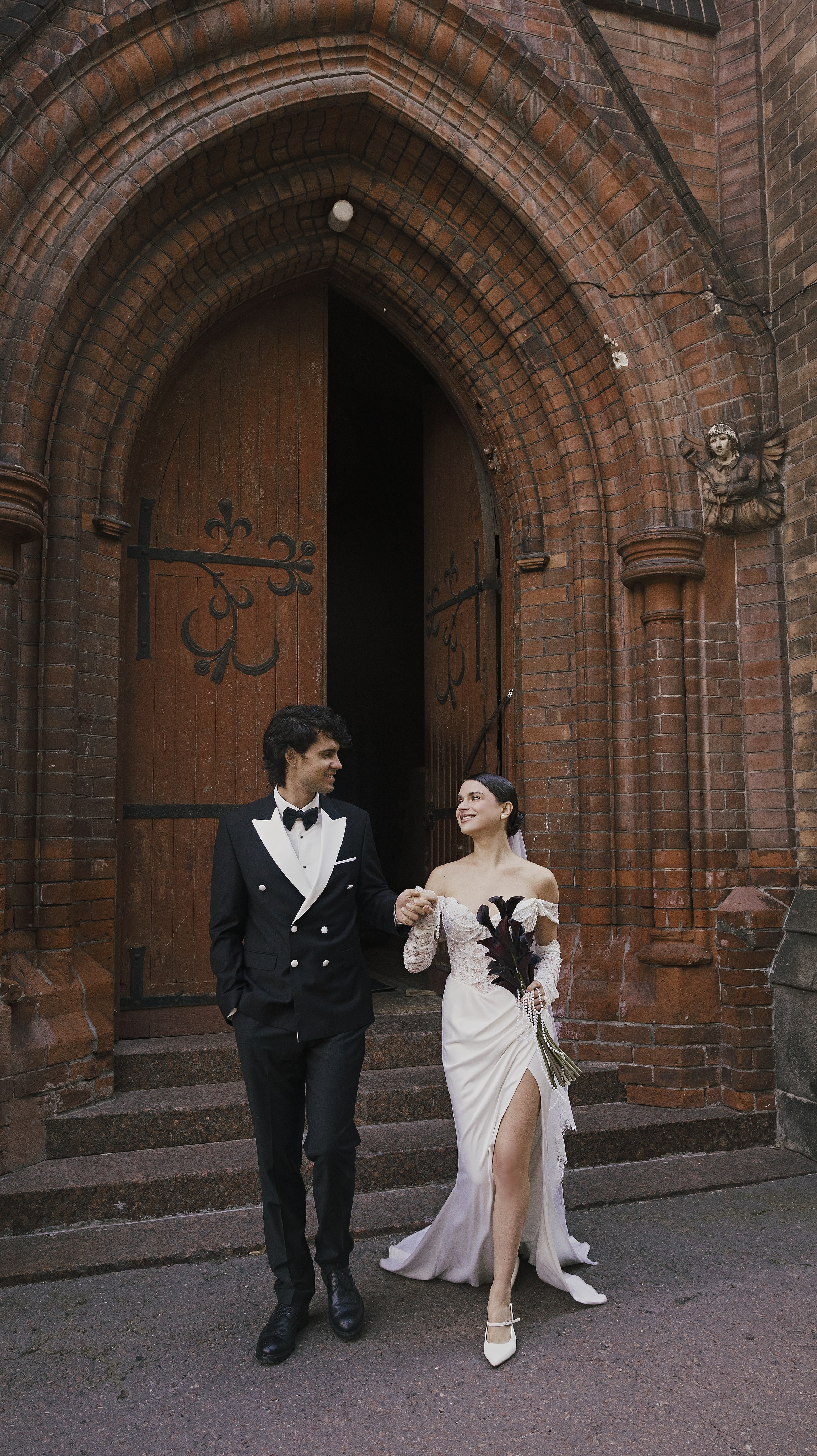 «Свадебная фотография в полный рост: невеста в платье с открытыми плечами и высоким разрезом и жених в двубортном смокинге, идущие рука об руку и улыбающиеся перед большой кирпичной церковью с арочным деревянным входом».