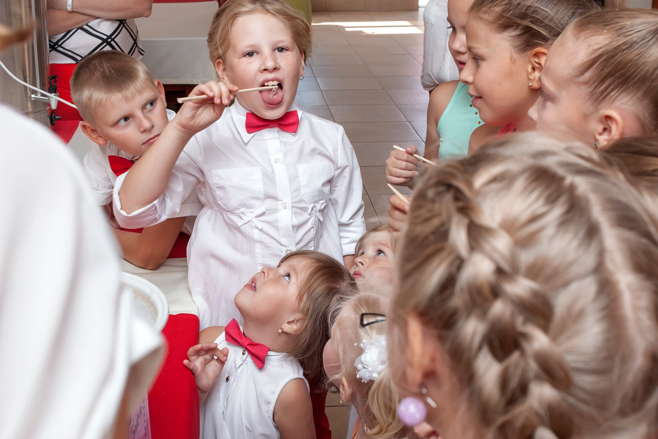 Девочка в белой рубашке пробует сахарную вату на палочке, остальные дети смотрят на неё. 