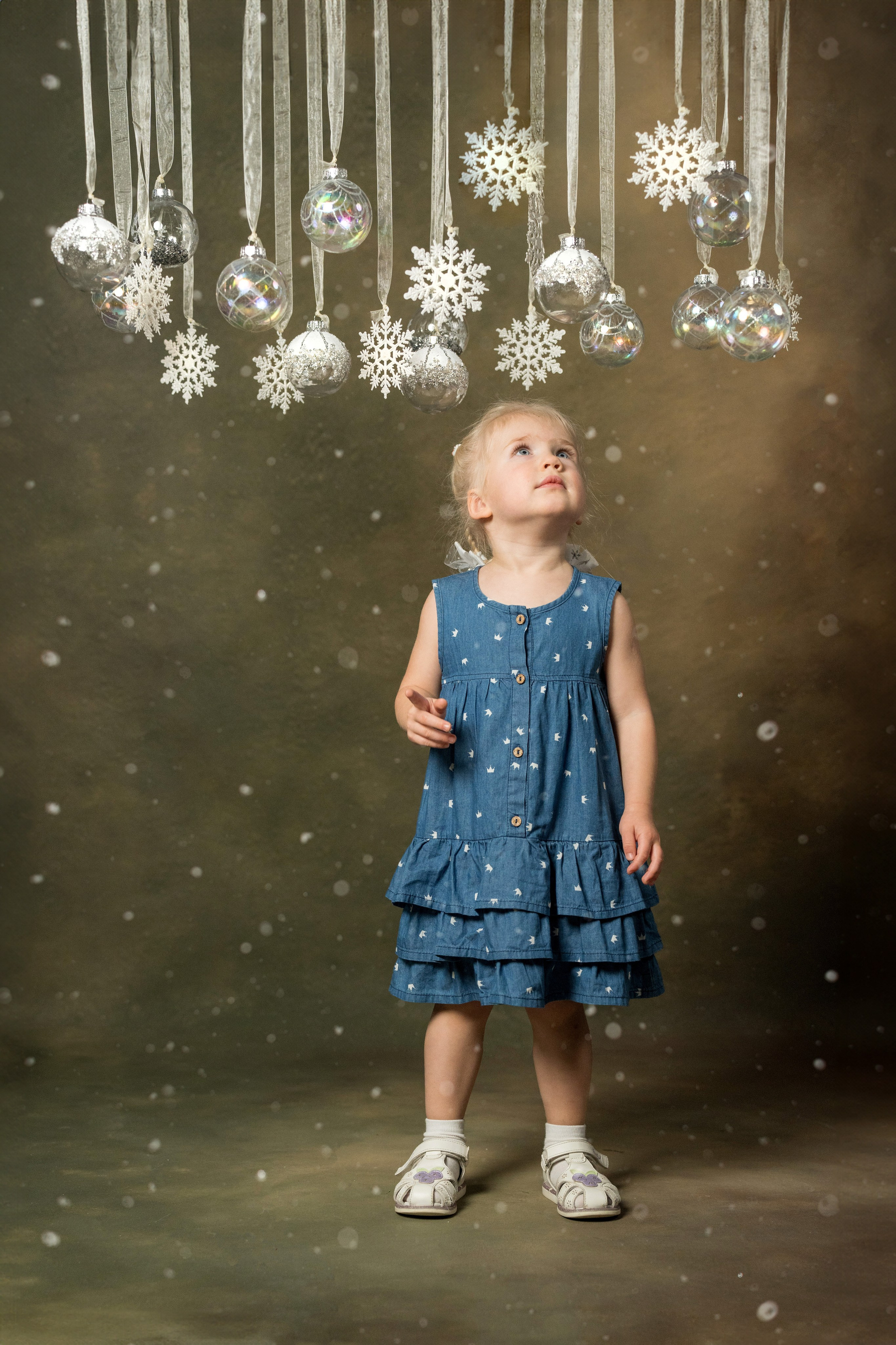 Малышка под новогодними шарами с интересом смотрит на них а вокруг кружится снег.