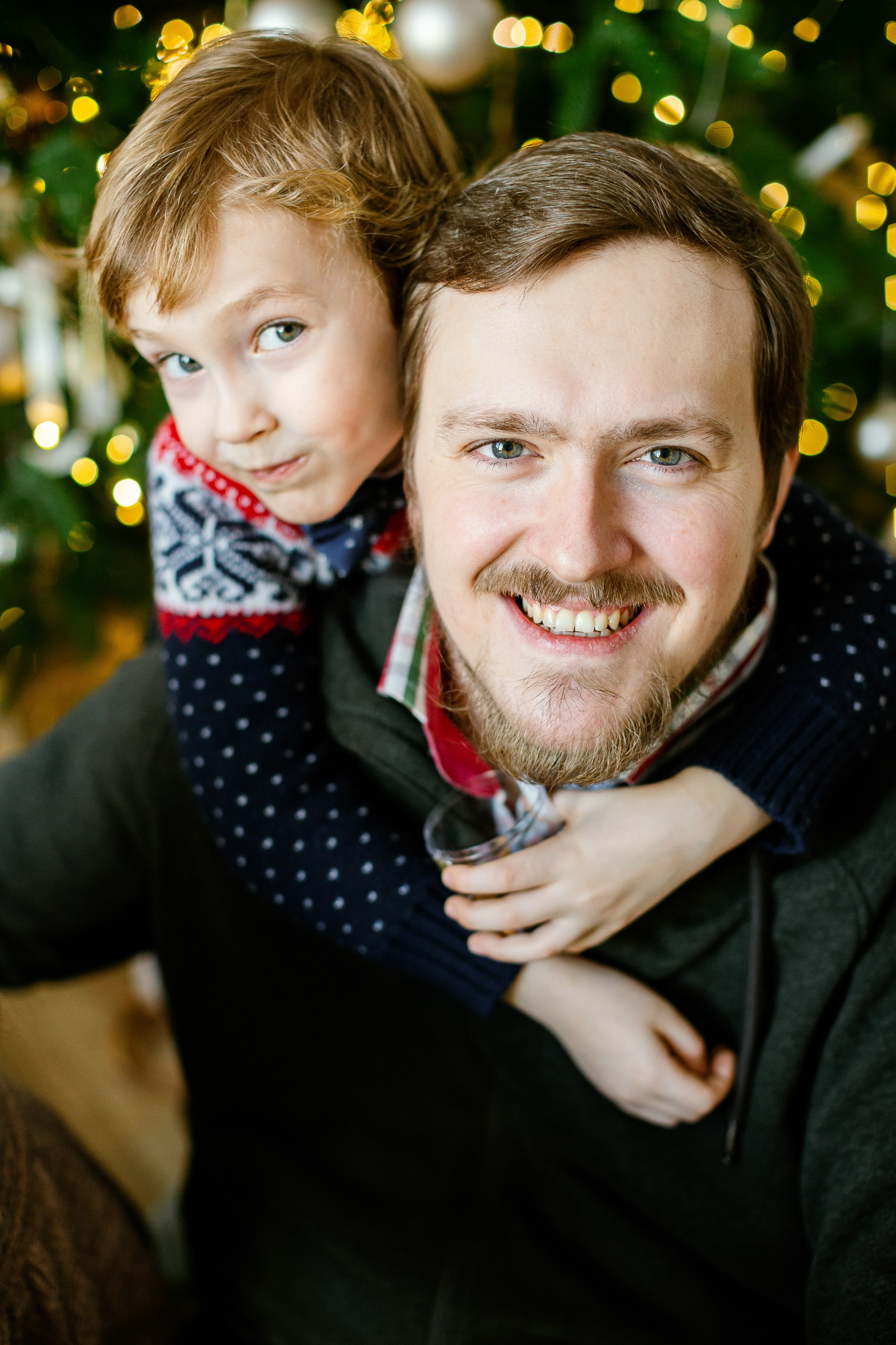 Папа и сын сидят на полу и обнимаются. На фоне стоит елка с огоньками.