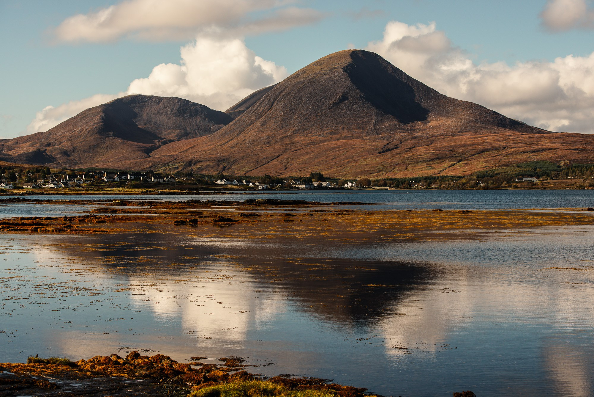 Суровый горный пейзаж острова Скай, Шотландия: большая коричневая гора отражается в спокойной воде залива, на берегу расположен ряд домов,