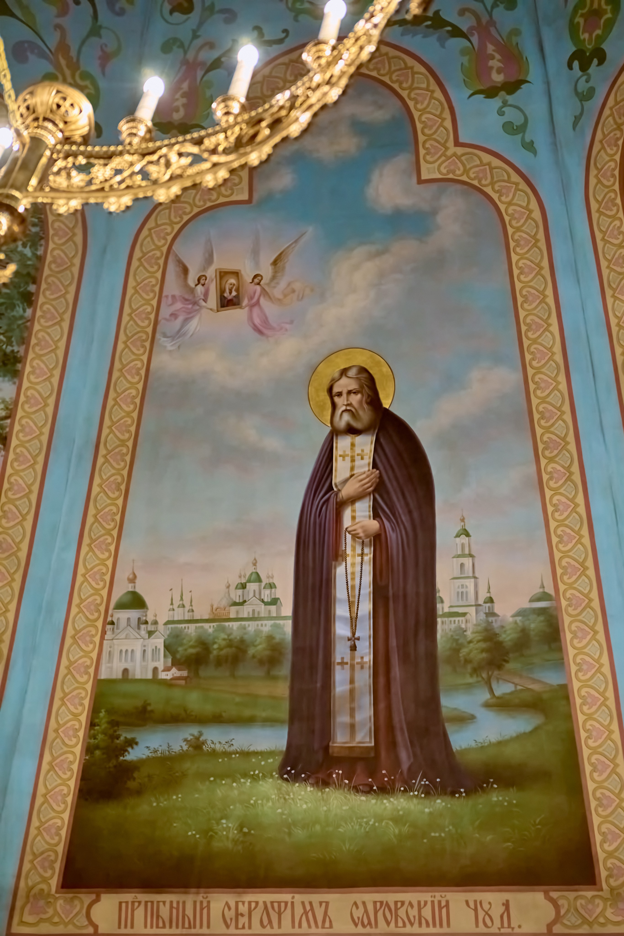 Фреска в полный рост Святаго Преподобного Серафима Саровского в храме. Преподобный стоит на возвышении на заднем плане Саровская обитель