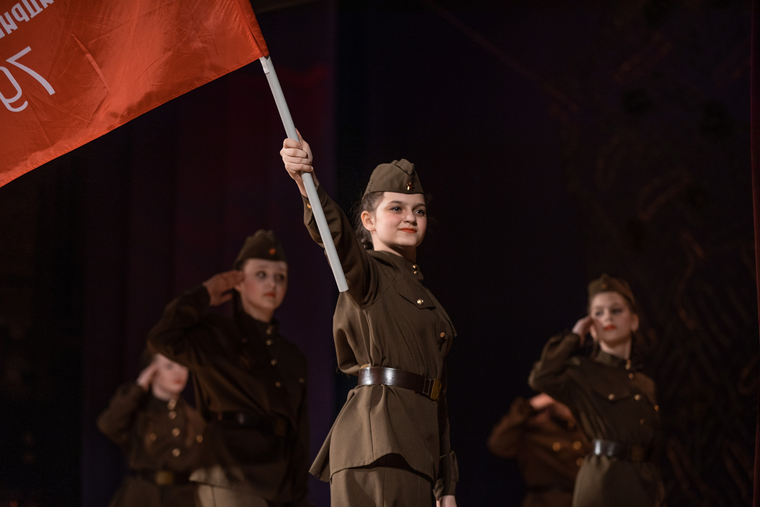 Подростковый коллектив выступает на концерте по случаю дня защитника Отечества. Девочка в форме с красным флагом на сцене.